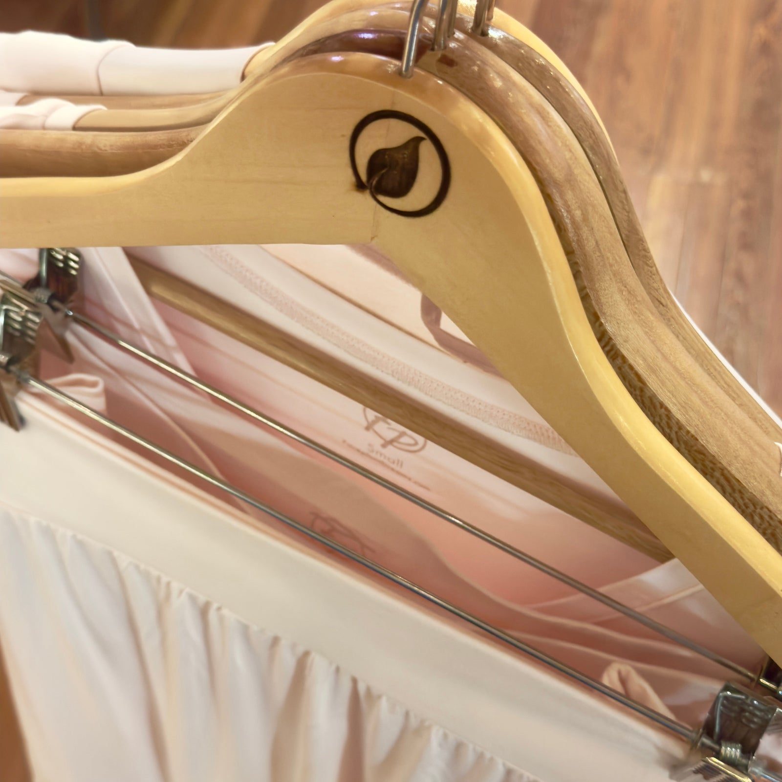Two wooden hangers with leaf logos hold light pink clothing on a metal rack. The hangers and clothes are neatly arranged, and the floor is wooden.
