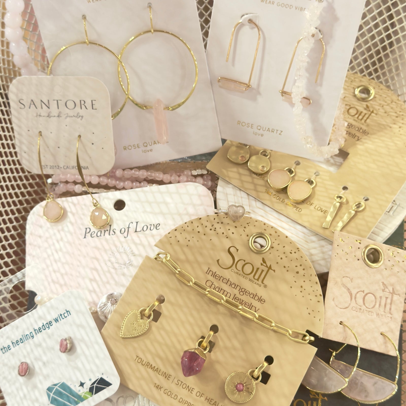 A collection of gold and rose quartz jewelry—including earrings, bracelets, and charms—displayed on branded cards with decorative backgrounds.