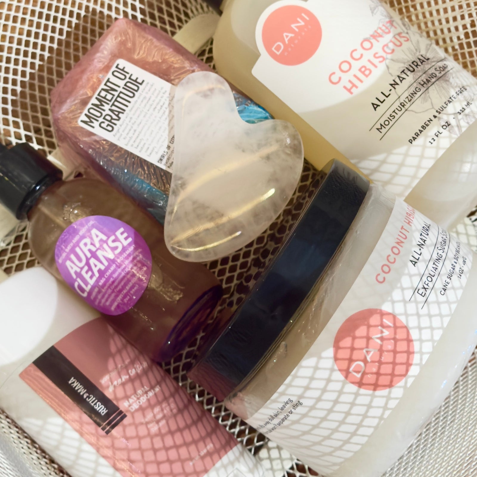 A basket containing self-care products including a bar soap, a crystal facial tool, lotion, exfoliating scrub, and spray labeled Aura Cleanse and Moment of Gratitude.