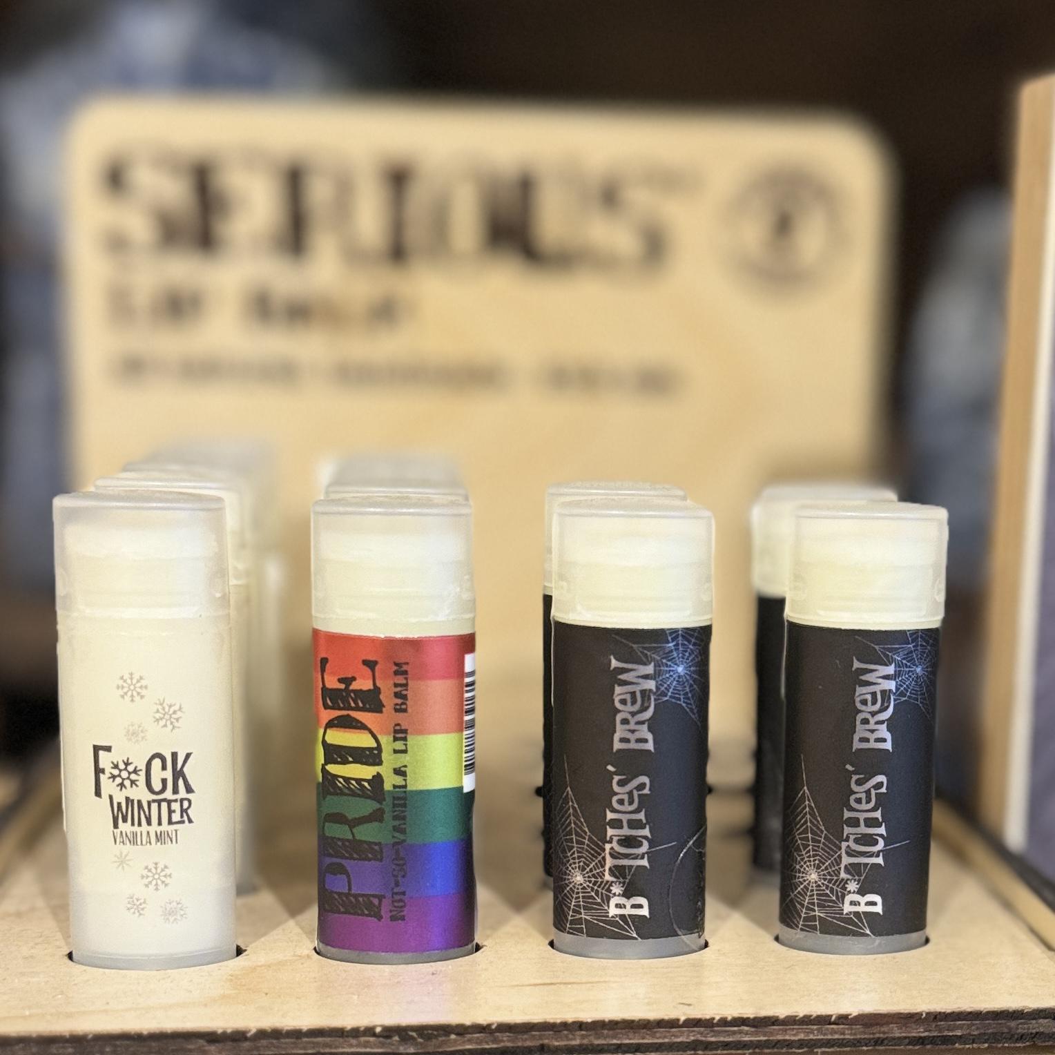 Four upright handmade Serious Boop Balm tubes by The Healing Hedge Witch, featuring unique labels like F*CK WINTER, PRIDE, and Blues Brew, are displayed on a shelf in Jacksonville, Illinois with a blurred logo in the background.