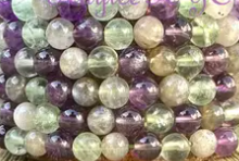 A close-up of The Healing Hedge Witch Throat Chakra Crystal Bracelet, featuring polished, shiny round beads in green, purple, and clear hues arranged in neat rows and resembling semi-precious stones.