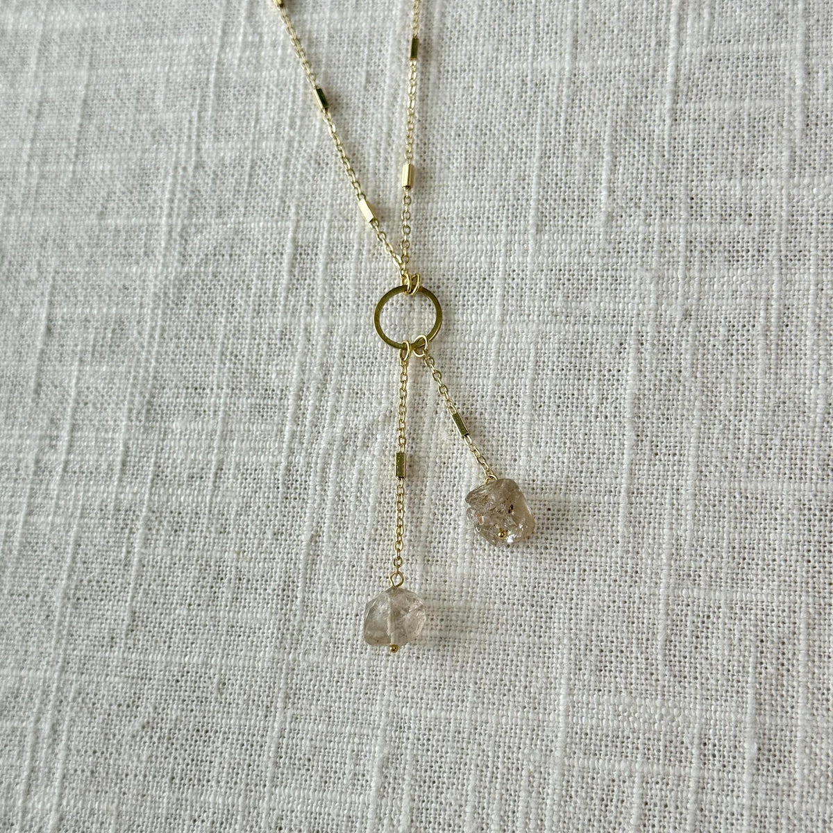 The Healing Hedge Witch’s Herkimer Diamond Tassel necklace features two chains ending with raw Herkimer Diamonds, centered by a delicate gold loop—ideal for meditation and awakening the Crown chakra, shown on textured white fabric.