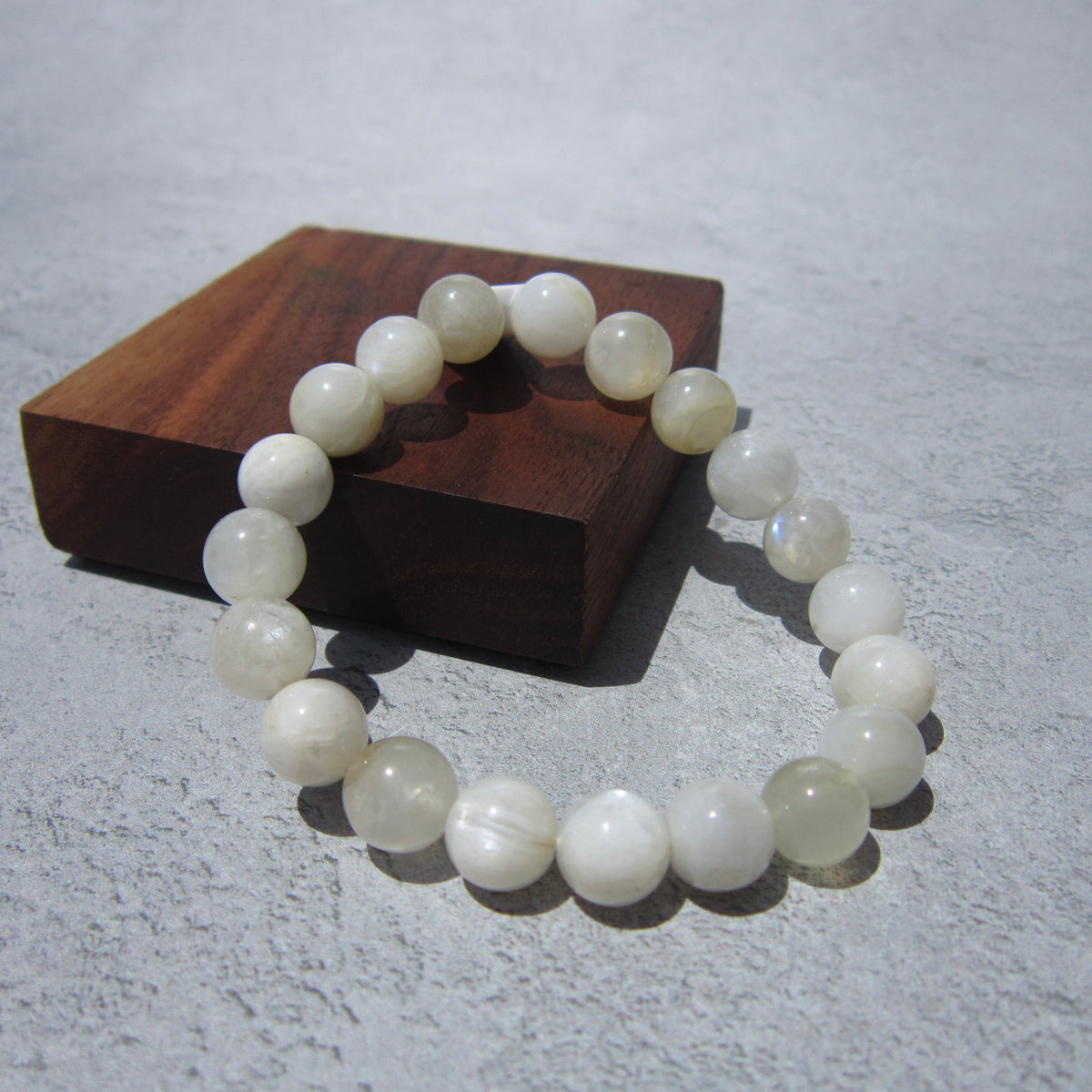 The Healing Hedge Witch Moonstone Bracelet, featuring round translucent white beads, is displayed on a square wooden block against a textured light gray background.