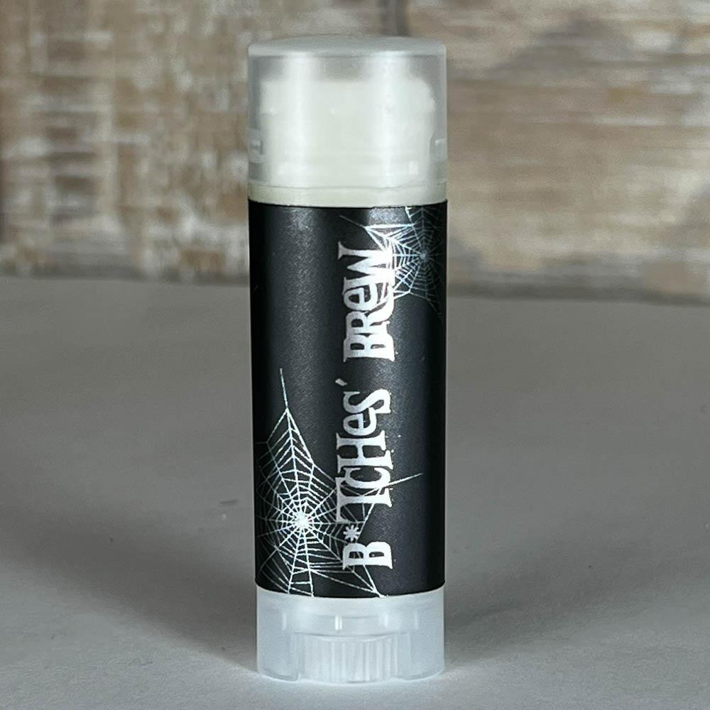 A tube of Serious Boop Balm by The Healing Hedge Witch with a black label and white spiderwebs stands on a gray surface, blurred wood background. Handmade in Jacksonville, Illinois with natural flavor oils.