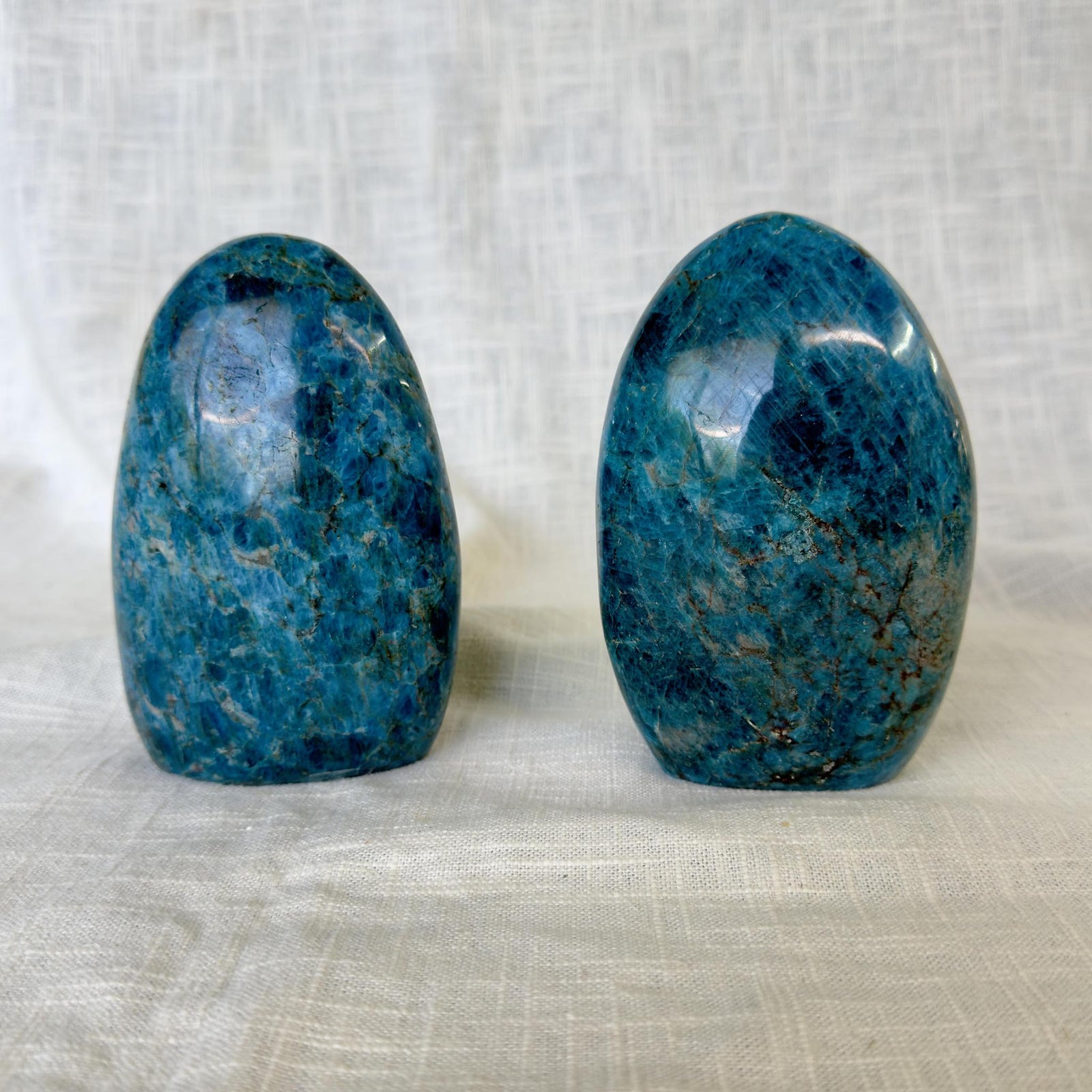 Two polished Blue Apatite Freeforms from The Healing Hedge Witch with rounded tops are displayed upright on white textured fabric, ideal for inspiring creativity and activating the Third Eye chakra.