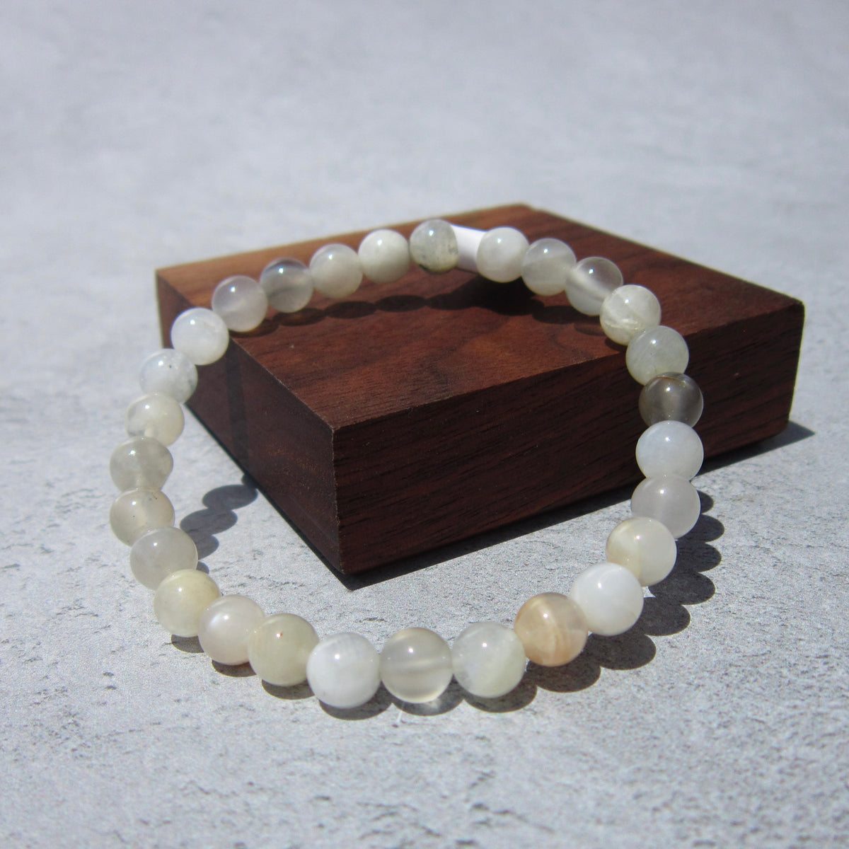 The Healing Hedge Witch Moonstone Bracelet, featuring round, translucent stones, is displayed on a gray surface with part of the bracelet resting on a small rectangular wooden block.