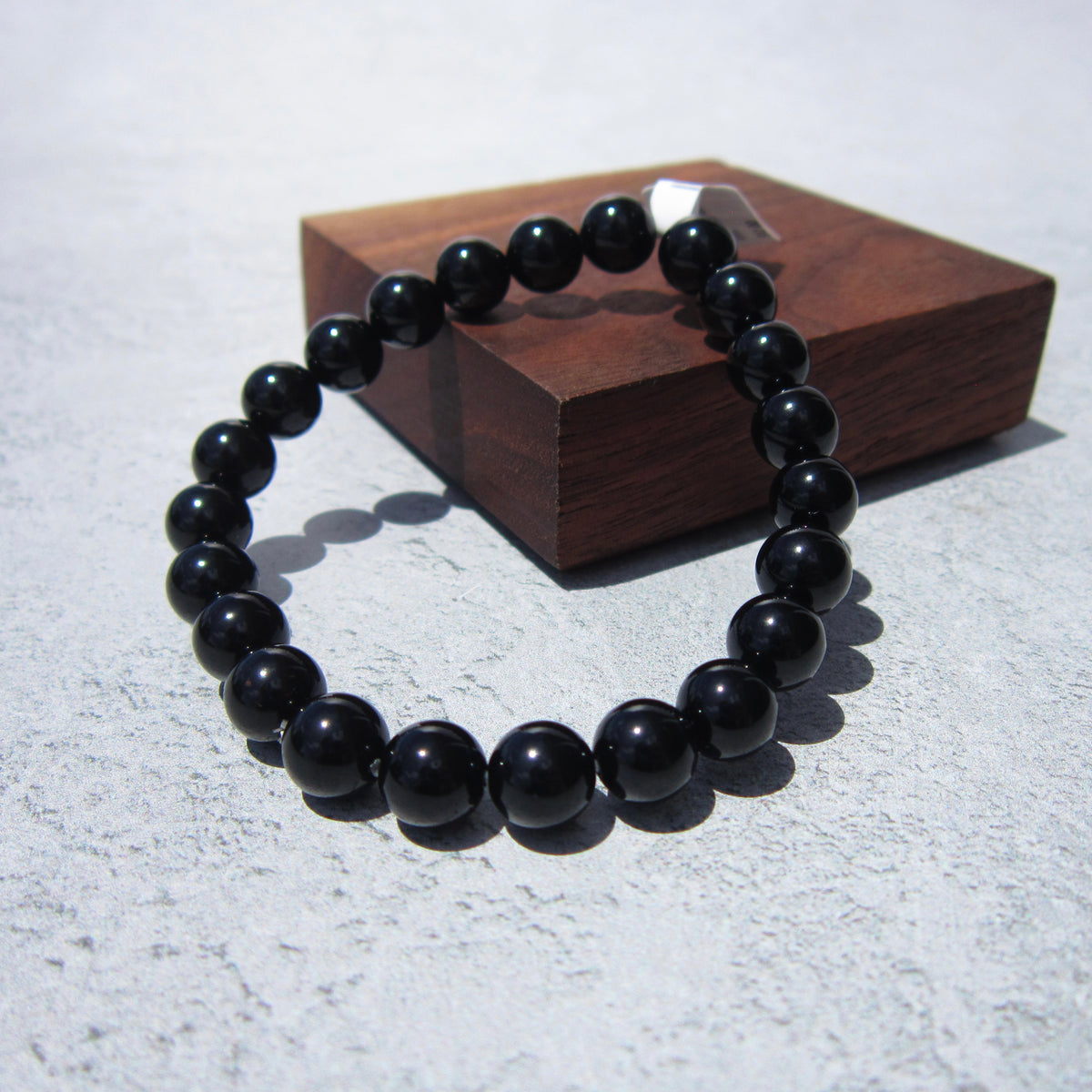 The Healing Hedge Witch Obsidian Bracelet, known for grounding and protection, is displayed in a circle on a textured light gray surface with a wooden box in the background.