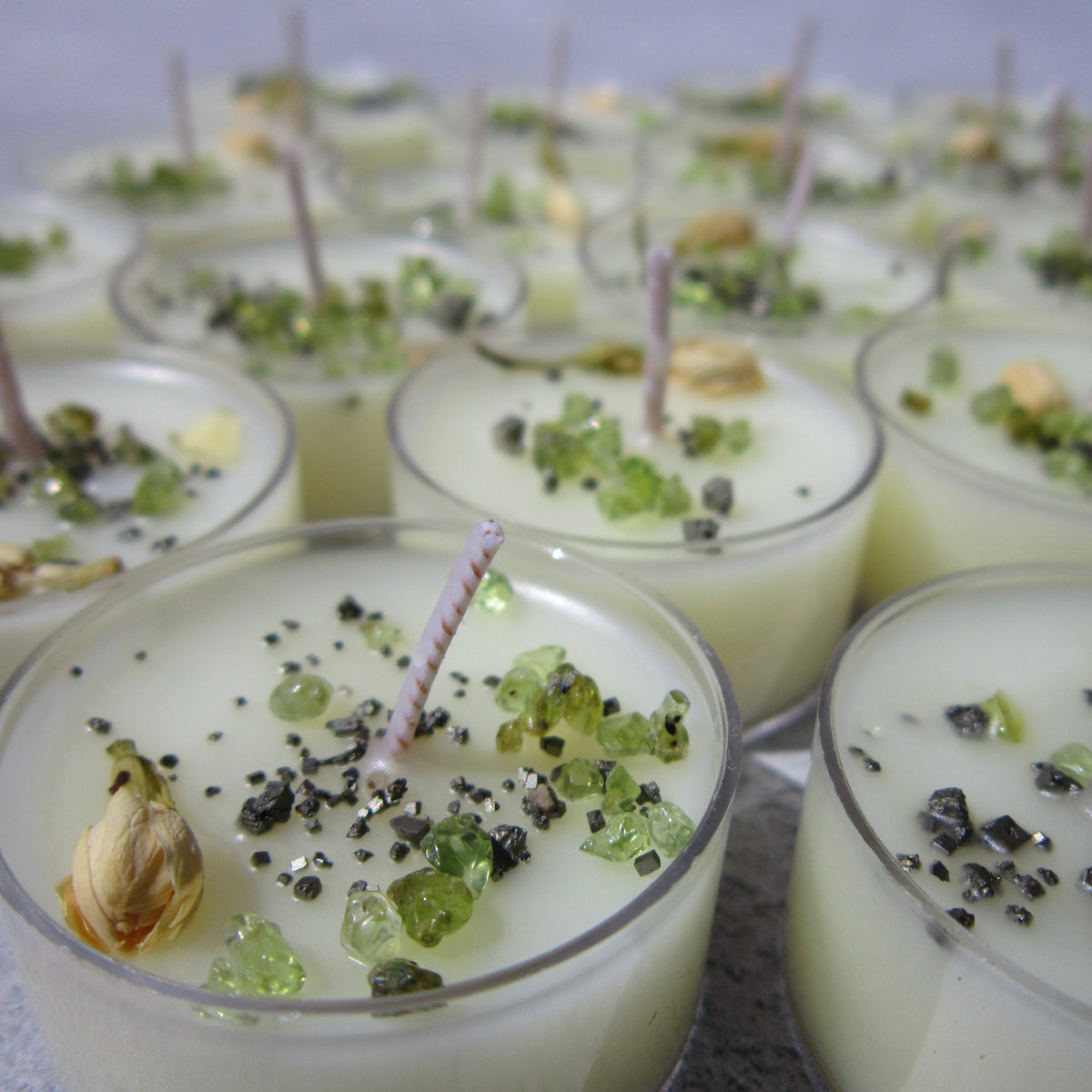 A close-up of The Healing Hedge Witch Money Maker Tealights—white candles in clear holders, each with twisted wicks and topped with green crystals, dried herbs, and black specks.