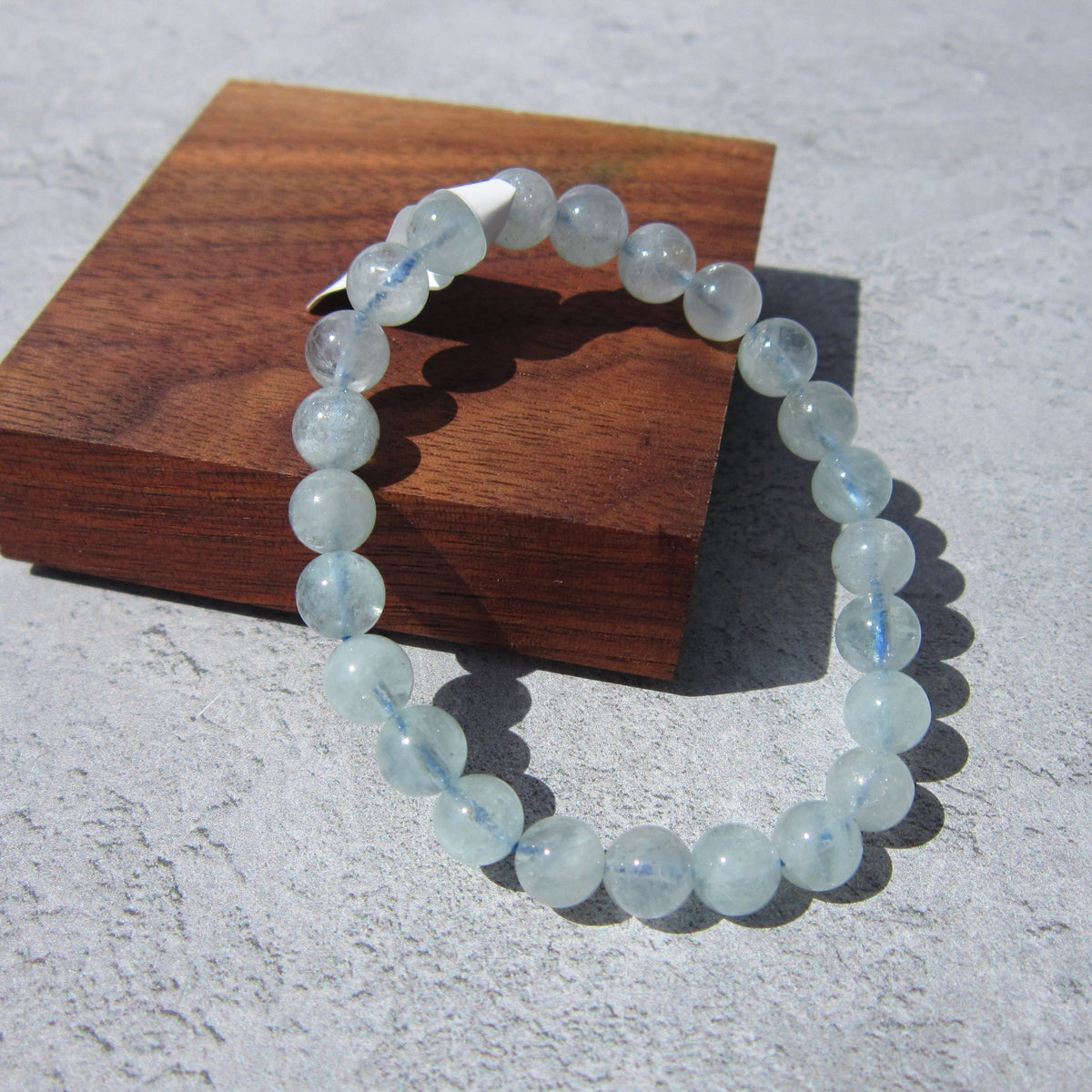 The Healing Hedge Witch's Aquamarine Crystal Bracelet, featuring round, light blue translucent beads, is shown on a gray surface with part of it resting on a wooden block.