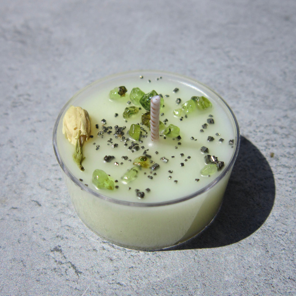 The Healing Hedge Witch’s Money Maker Tealight—a small, pale green candle in a clear holder with green crystal chips, black specks, and a dried white flower—rests on a gray surface.