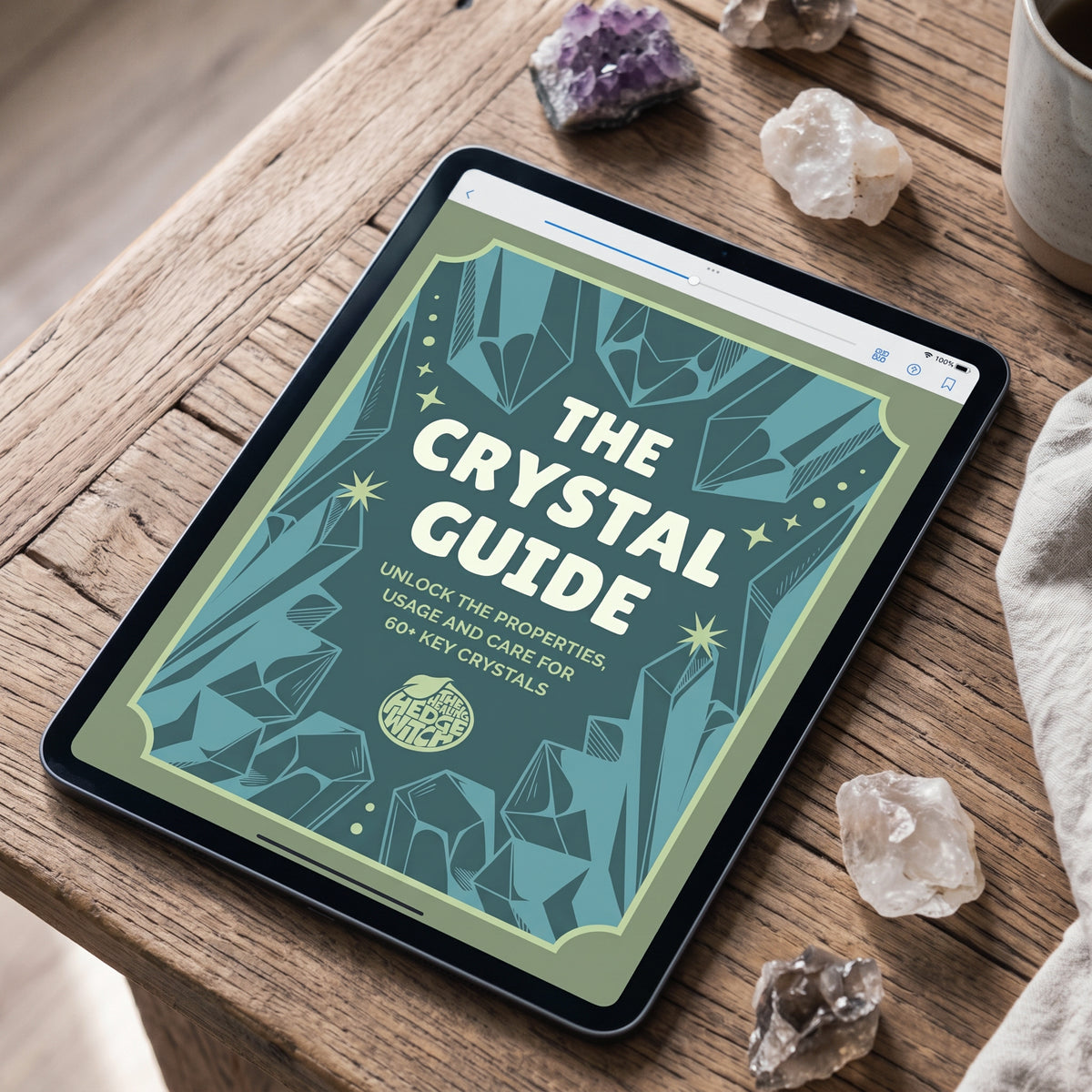 A tablet on a wooden surface displaying an ebook cover titled "The Crystal Guide, Unlock the Properties, Usage and Care for 60+ Key Crystals" surrounded by crystal stones.