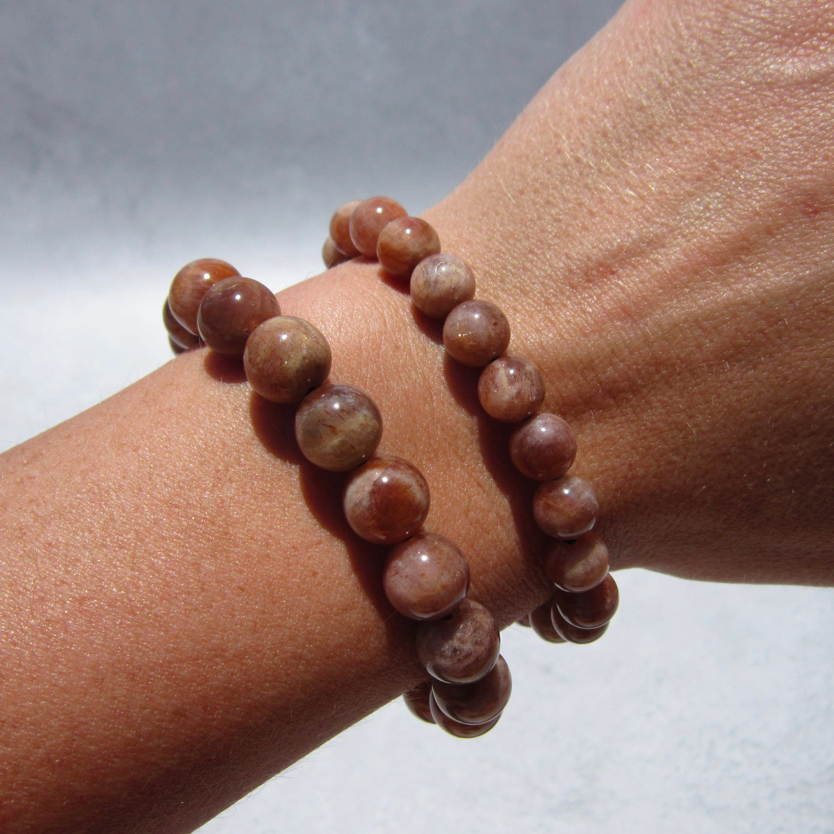 A close-up of a wrist wearing two Sunstone Crystal Bracelets by The Healing Hedge Witch, featuring polished brown stones believed to radiate healing energy, against a light background.