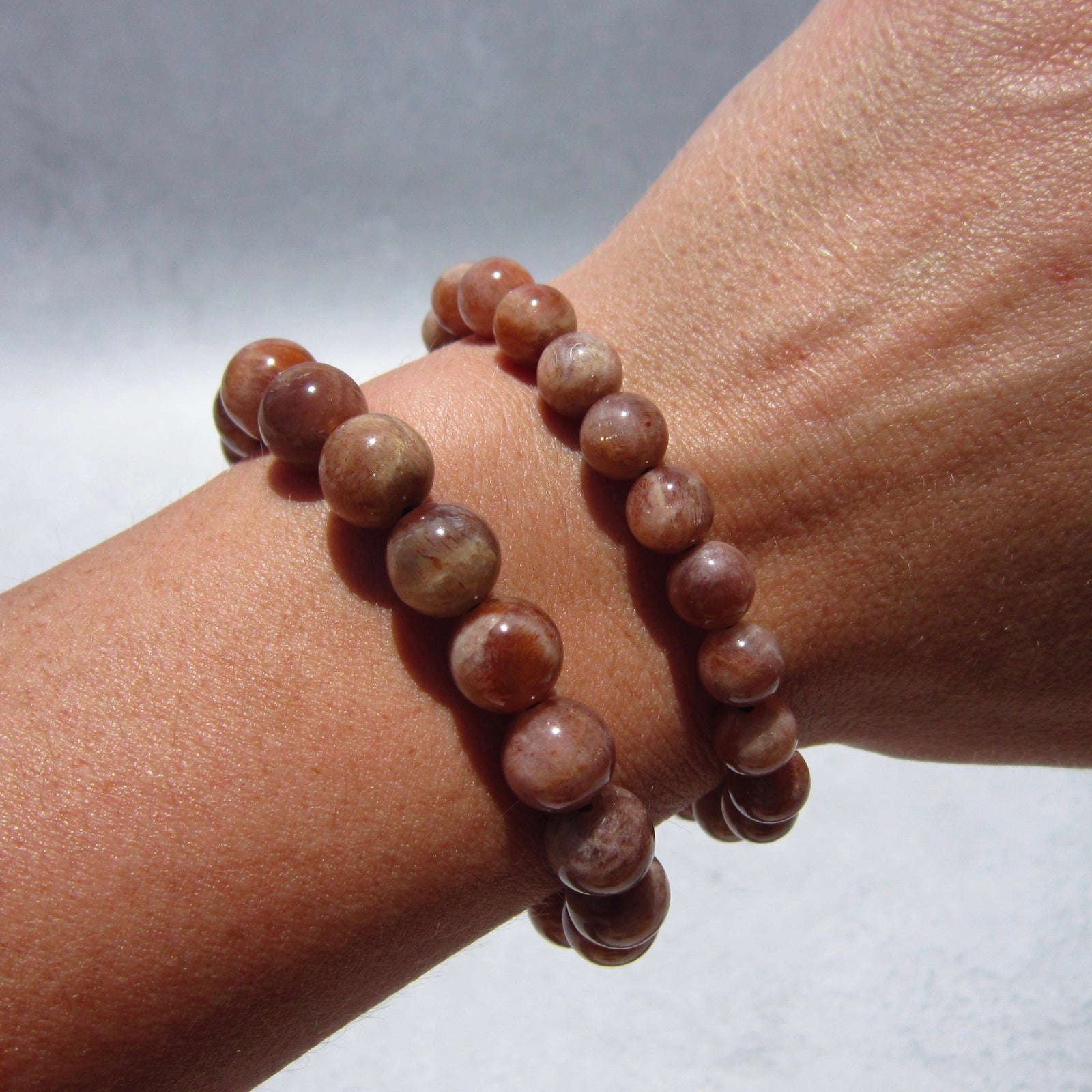 A close-up of a wrist wearing two Sunstone Crystal Bracelets by The Healing Hedge Witch, featuring polished brown stones believed to radiate healing energy, against a light background.