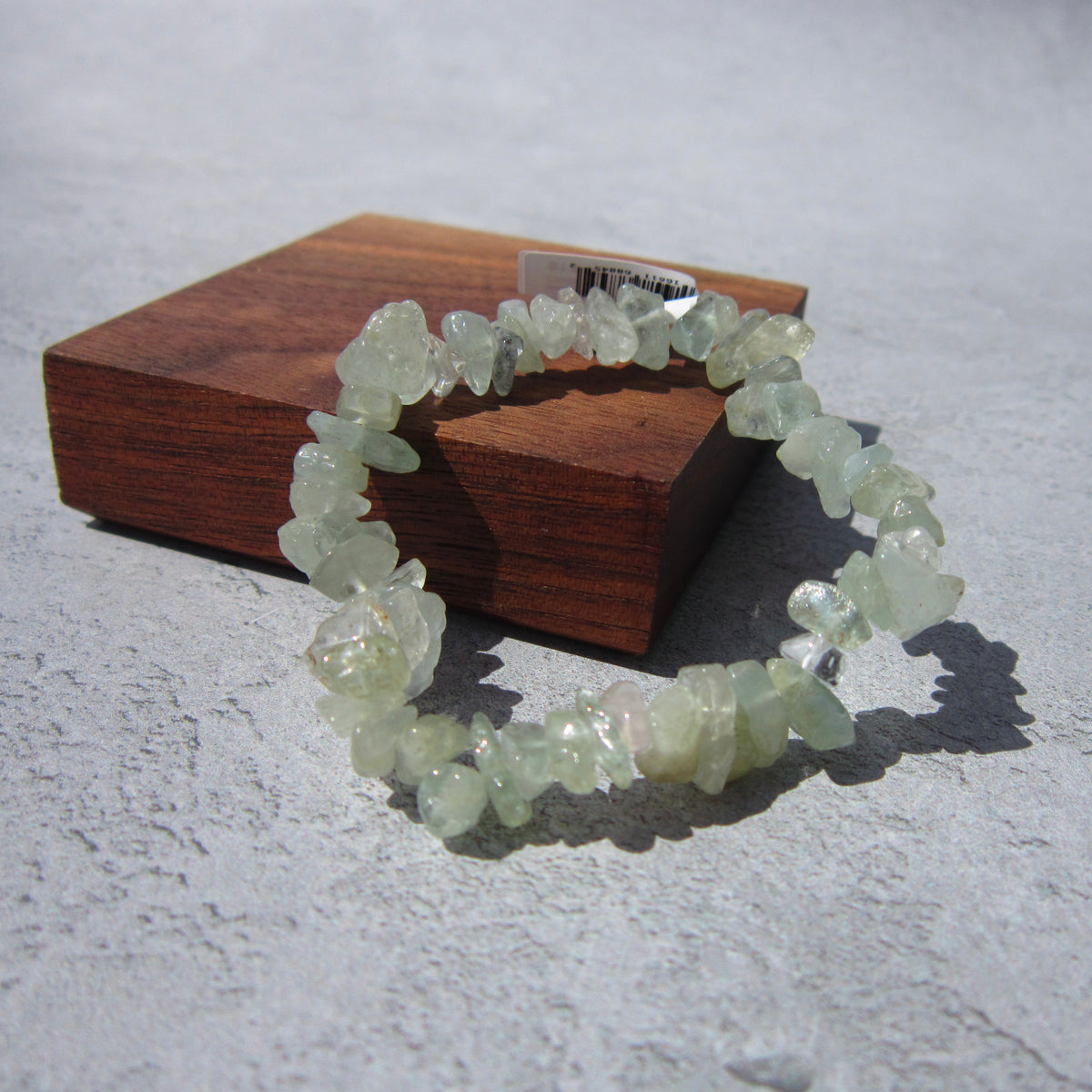 The Healing Hedge Witch Aquamarine Crystal Bracelet, featuring pale green gemstone chips, rests on a wooden block against a light gray textured surface with a small white barcode sticker visible.