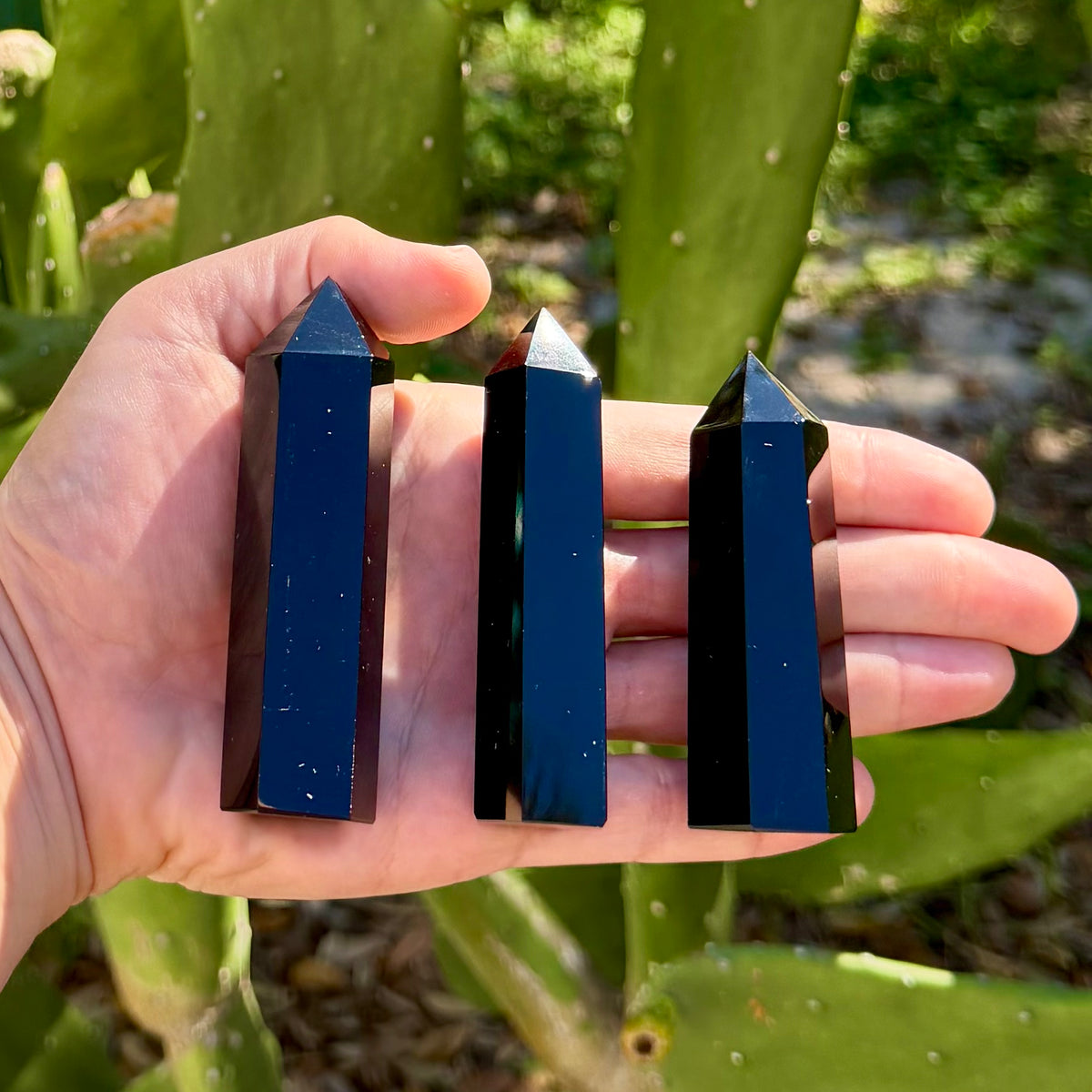 Three polished black obsidian crystal towers with faceted pointed tips and glossy rectangular column bodies, held upright in an open palm to show their size, set against an outdoor background of green cactus pads.