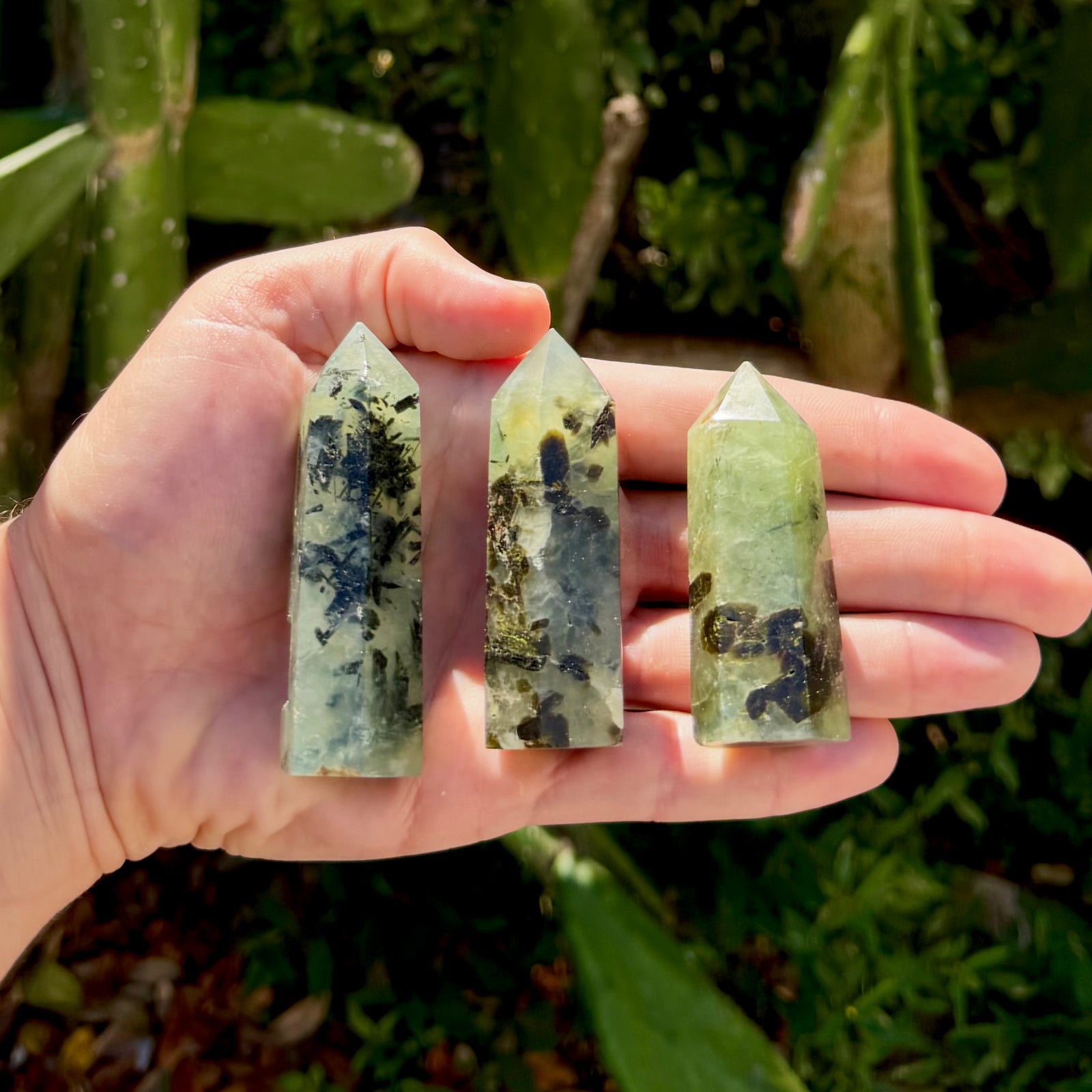 Three prehnite tower crystals held in an open palm outdoors against green foliage. Each is a polished pale green translucent column with a pointed top, marked with dark green to black inclusions. The three towers vary slightly in size and pattern.