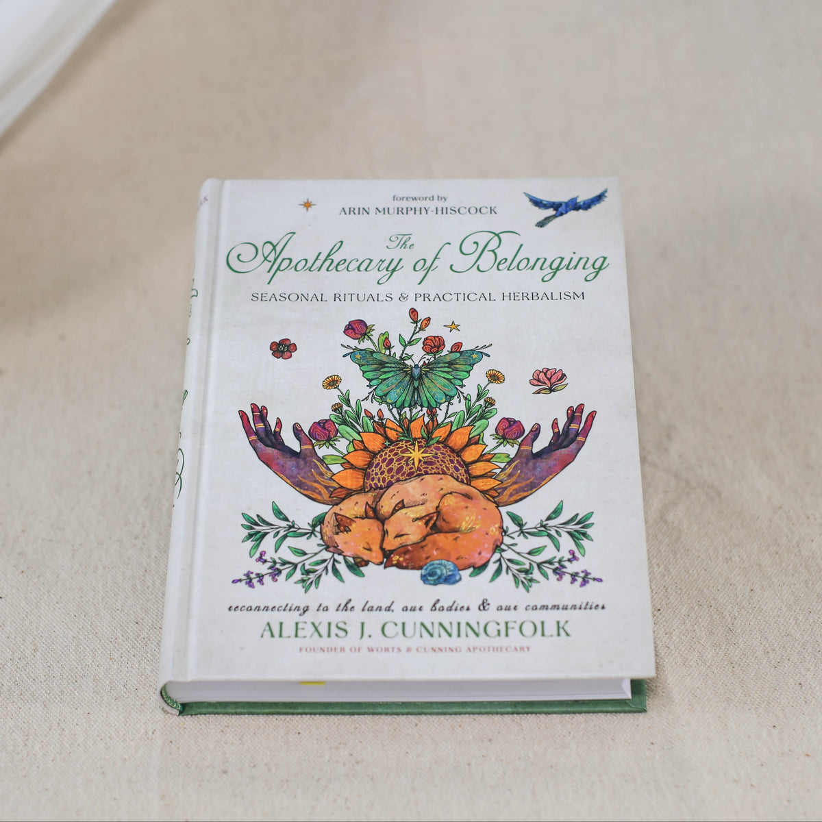 Apothecary Of Belonging by The Healing Hedge Witch rests on a beige surface, its cover adorned with vibrant botanical illustrations, hands holding flowers, and a bird perched at the top.