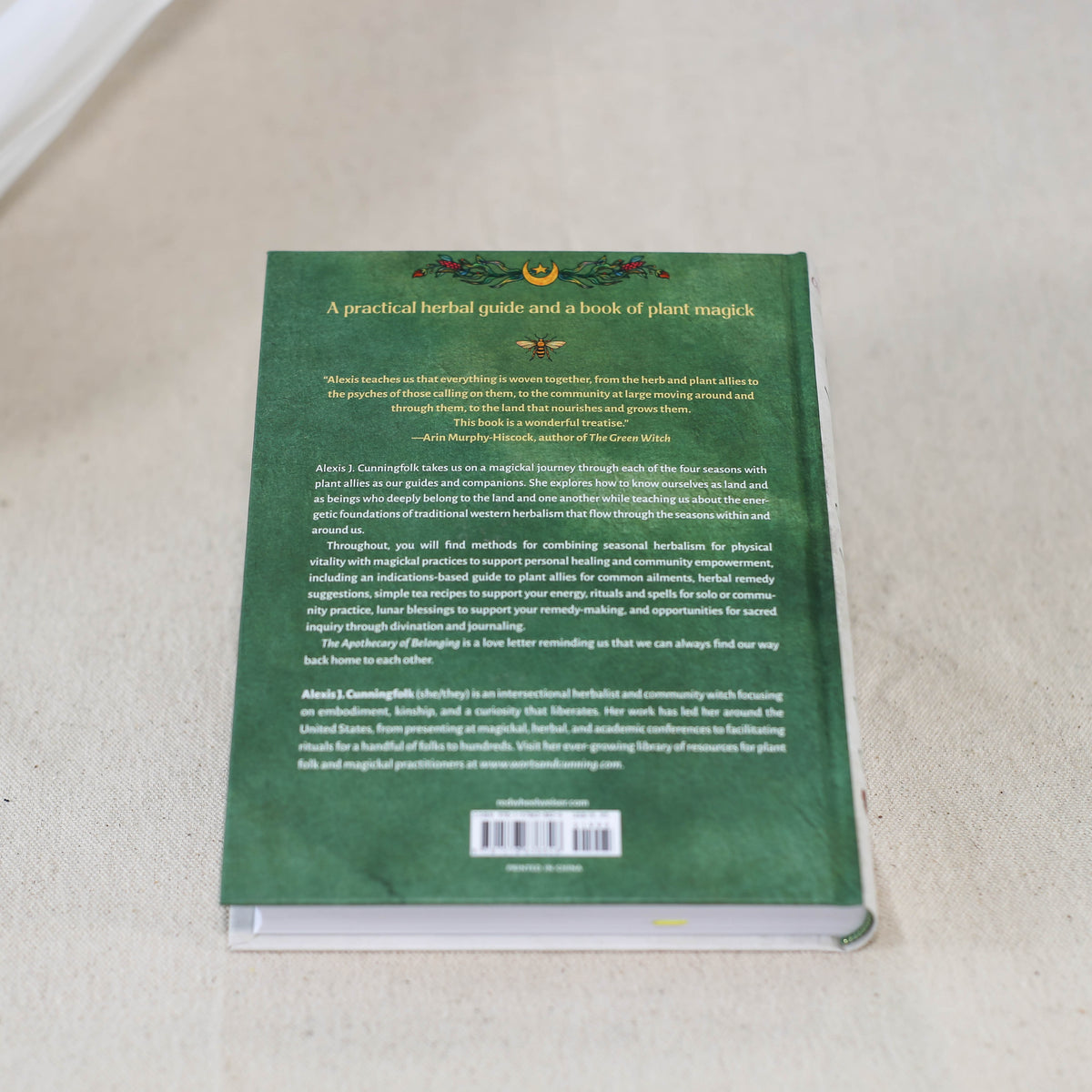 The back cover of a green hardcover book titled "Apothecary Of Belonging" by The Healing Hedge Witch, showcasing a summary and author bio, rests on a light-colored fabric surface.