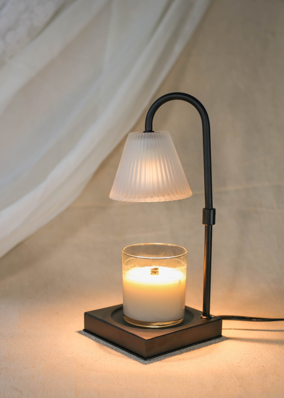 The Healing Hedge Witch's Lumiere Candle Warmer Lamp features a lit candle in a glass on a square black base, with a curved metal stem and ribbed glass shade, casting a warm glow against neutral fabric.