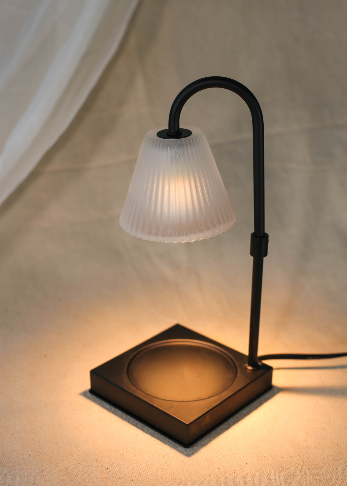 The Healing Hedge Witch’s Lumiere Candle Warmer Lamp features a modern black design with a frosted, ribbed glass shade. Switched on, it casts warm light over a beige fabric surface. The lamp has a square base and curved neck.