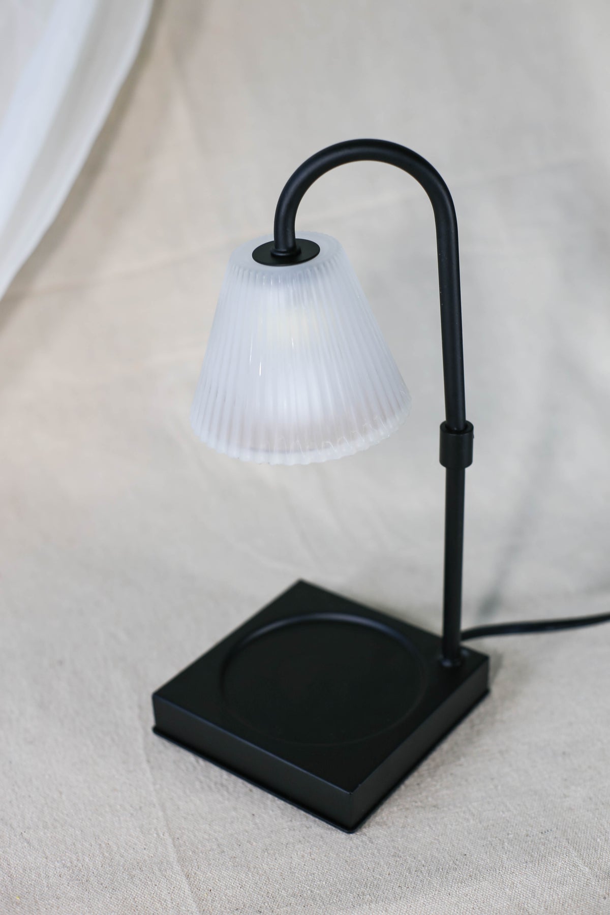 The Healing Hedge Witch's Lumiere Candle Warmer Lamp features a modern black base and ribbed white shade, showcased on a light fabric surface.