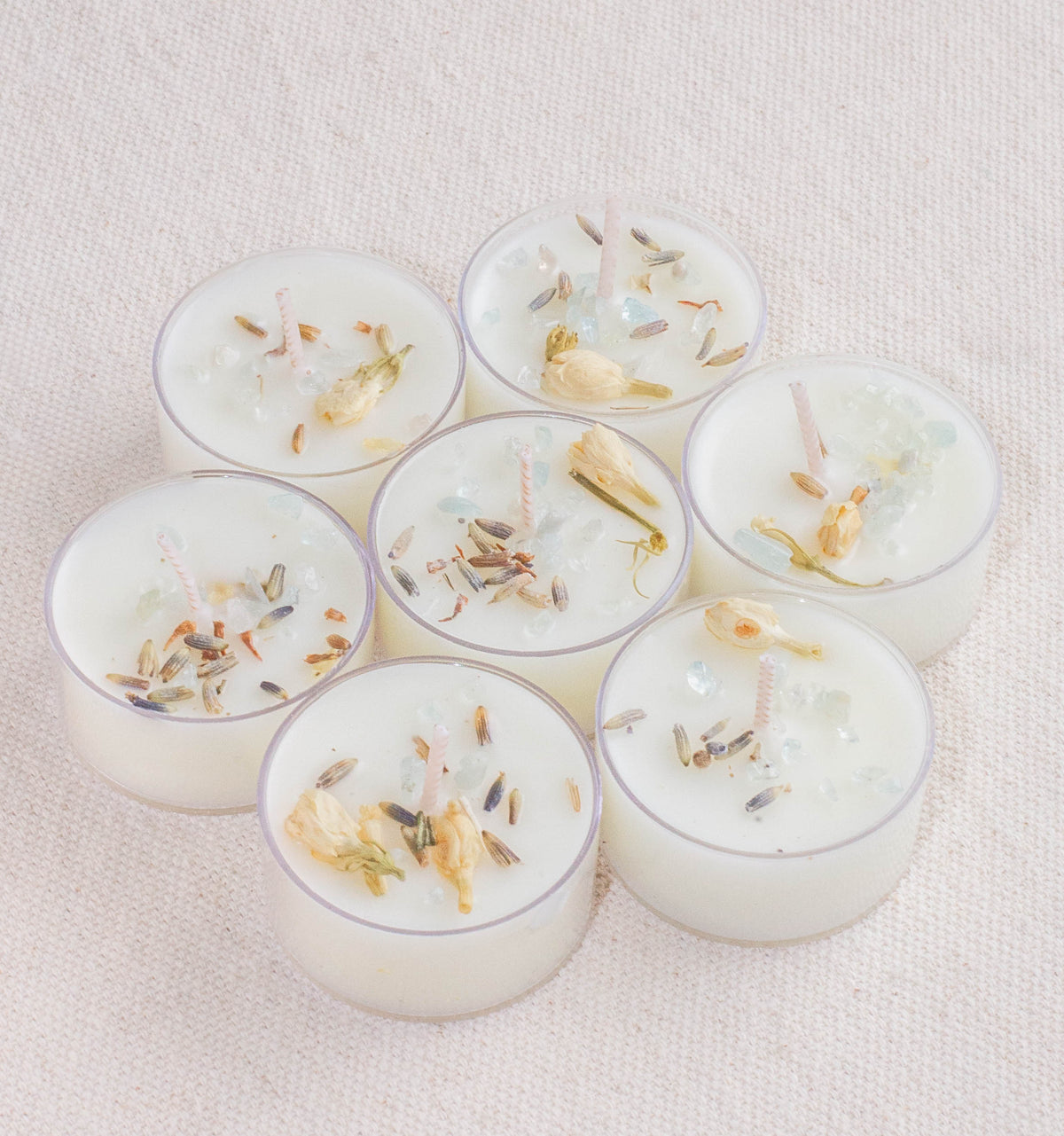 Seven Spring Equinox Spell Tealights by The Healing Hedge Witch are arranged in a circle on a light fabric, each one adorned with dried flowers, herbs, and tiny decorative crystals.