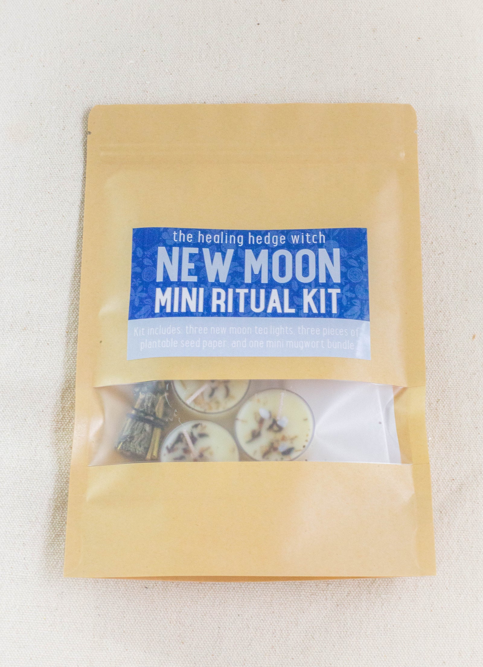 A brown paper pouch from The Healing Hedge Witch labeled New Moon Mini Ritual Kit features a blue and white sticker. Through its clear window, you see three tealights, a mini mugwort bundle, and plantable seed paper inside.