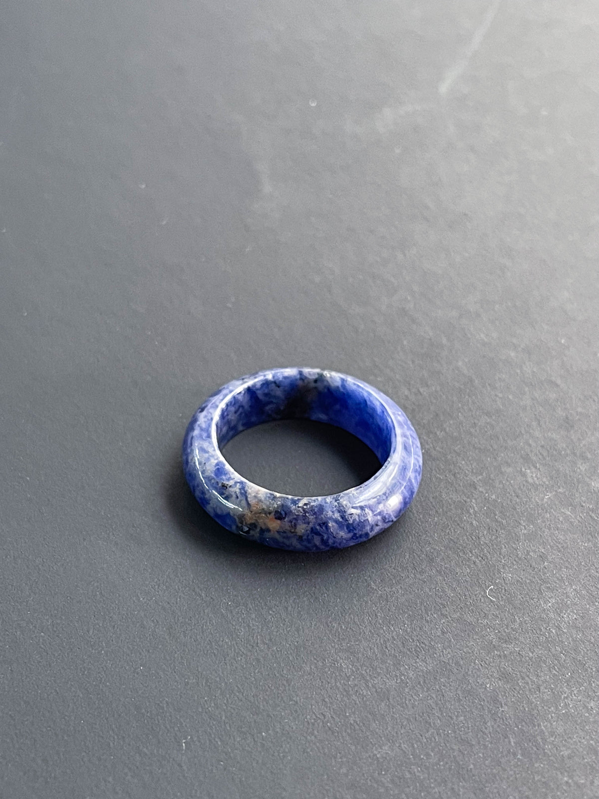The Healing Hedge Witch Sodalite Solid Ring, featuring blue and white marbled stone, rests on a smooth dark gray surface.