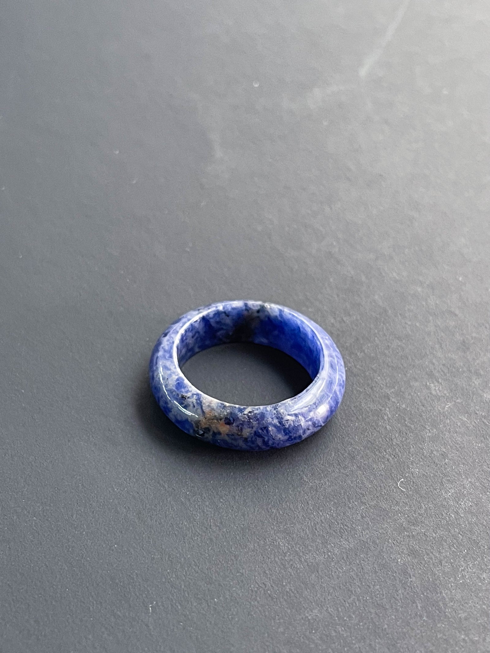 The Healing Hedge Witch Sodalite Solid Ring, featuring blue and white marbled stone, rests on a smooth dark gray surface.