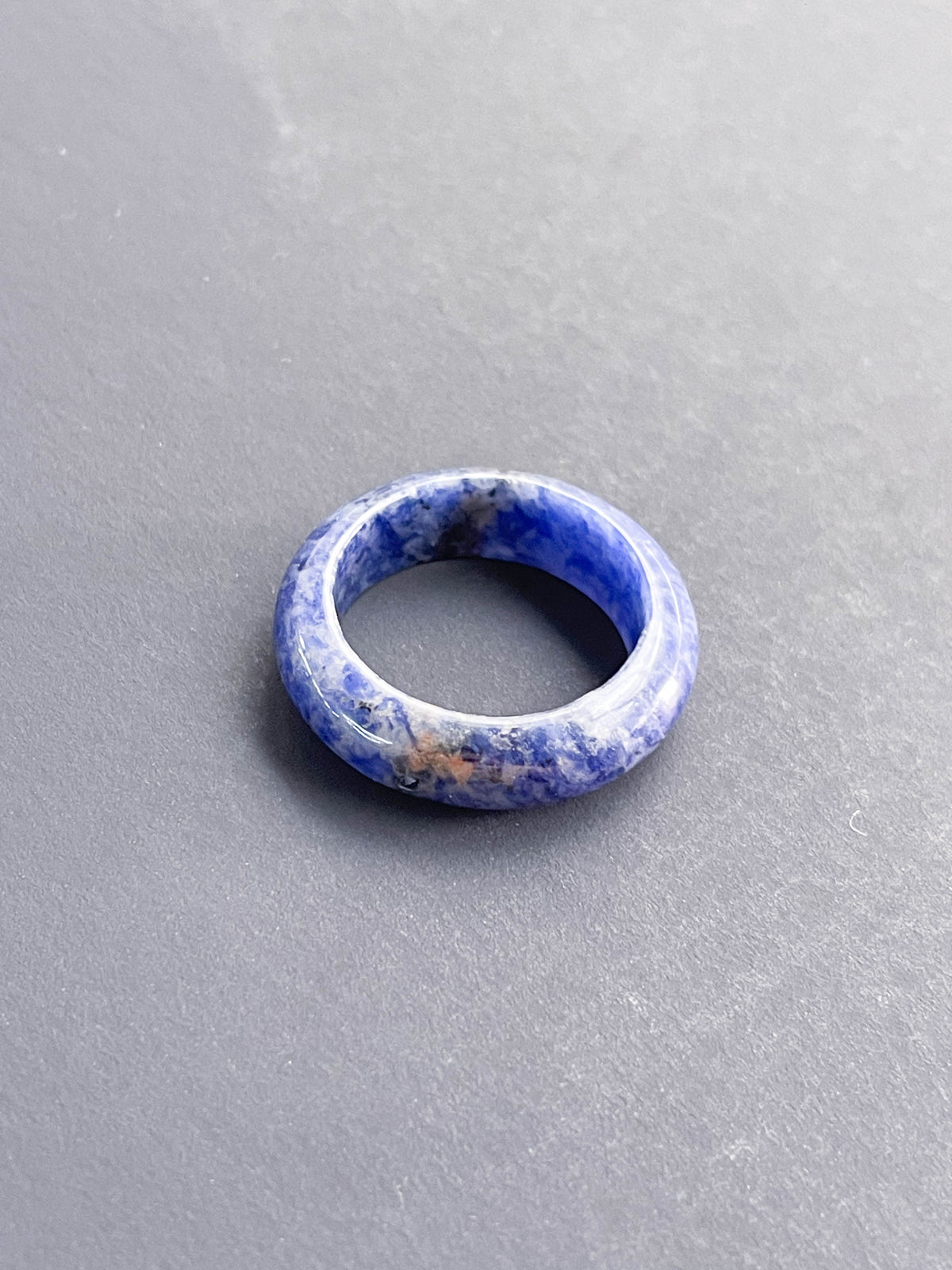 The Healing Hedge Witch's Sodalite Solid Ring, featuring blue marbled stone with white and light brown streaks, rests on a smooth gray surface.