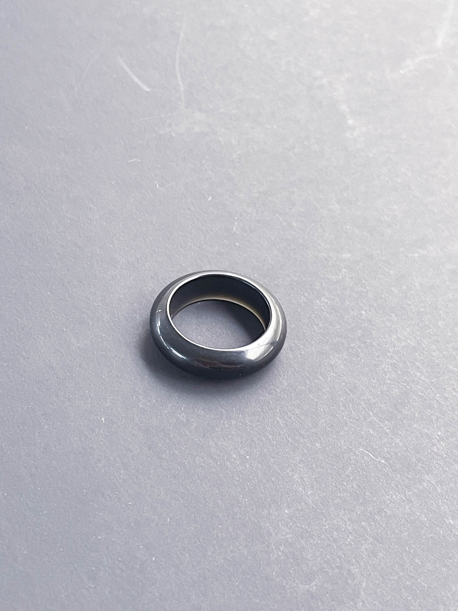 The Obsidian Solid Ring by The Healing Hedge Witch, featuring a shiny black finish and rounded edge, rests on a smooth, light gray surface.