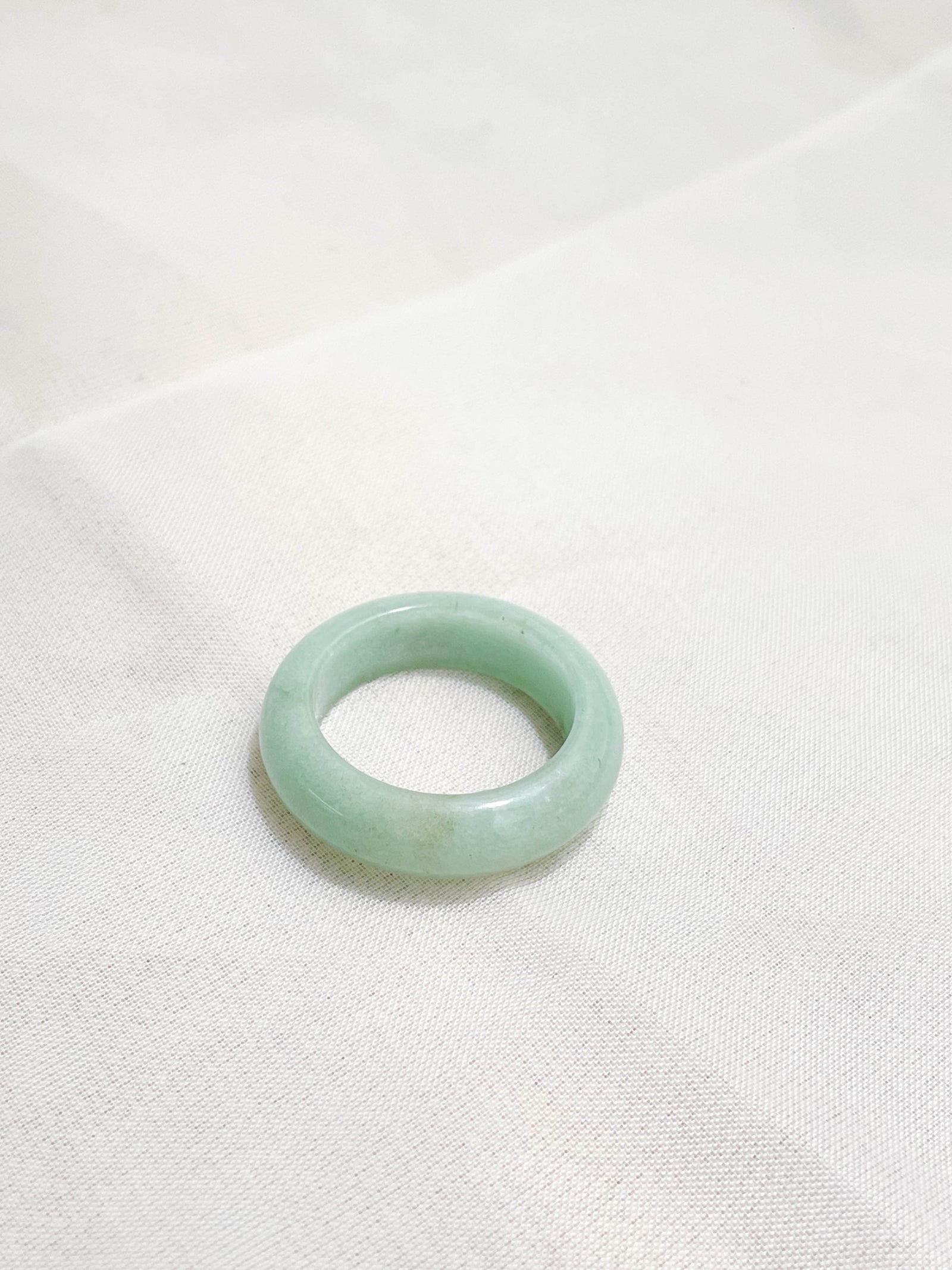 A pale green jade ring with a smooth, polished surface is placed on a white fabric background.