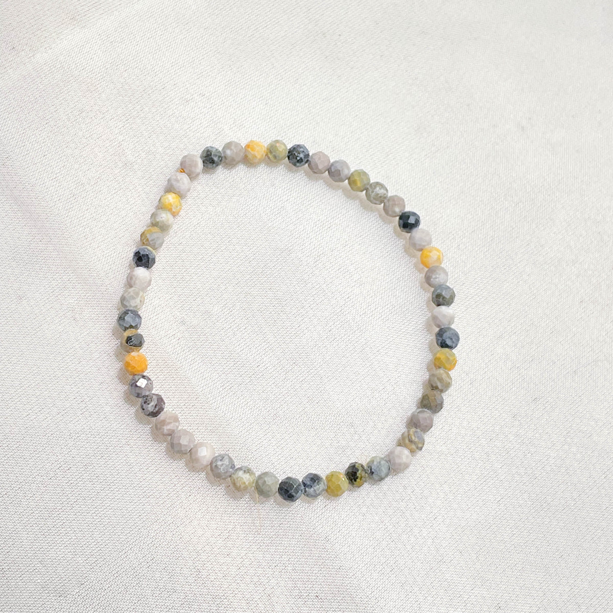 The Healing Hedge Witch’s Bumblebee Jasper Crystal Bracelet features delicate, faceted round beads in gray, white, yellow, and black on a single strand, beautifully displayed on a white fabric background.