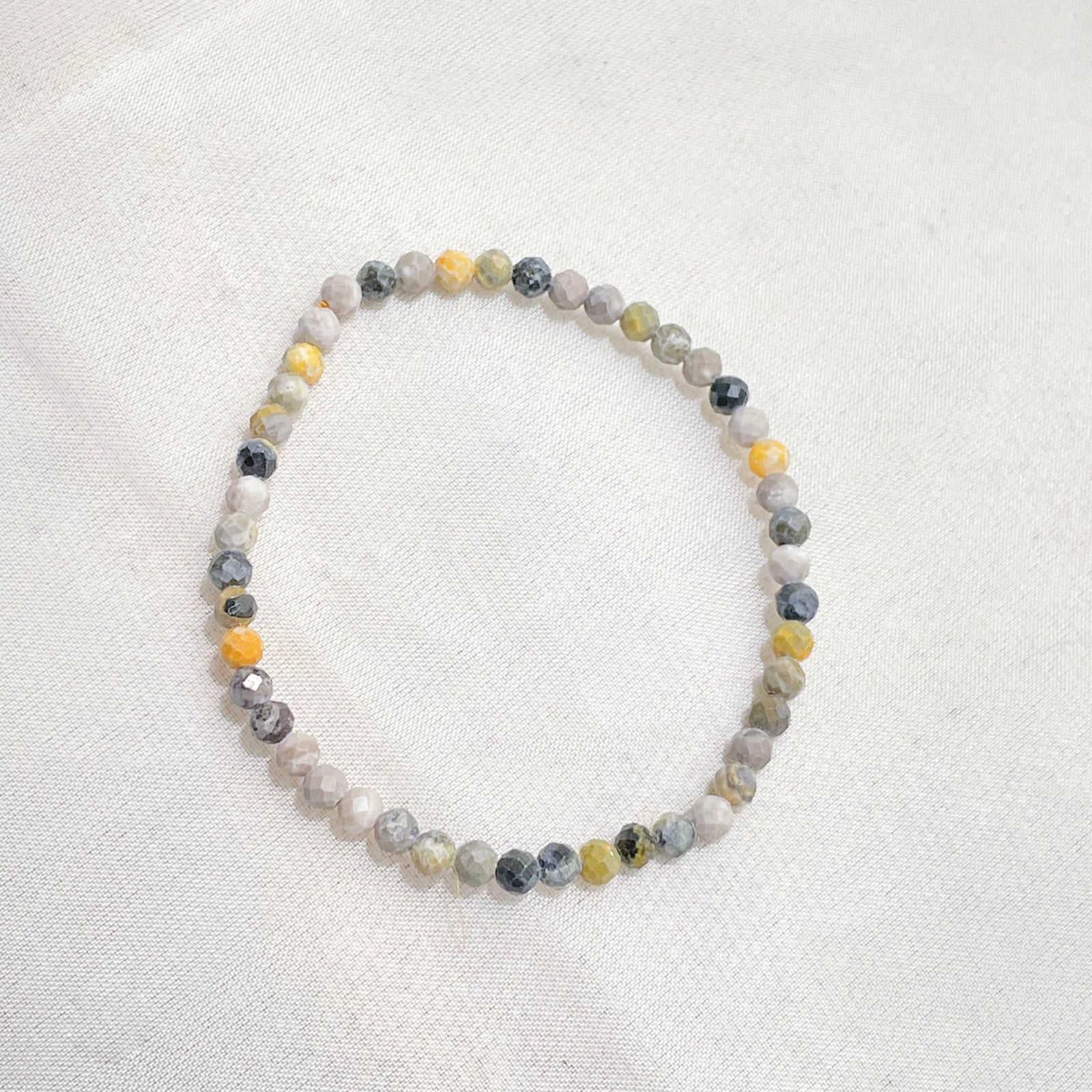 The Healing Hedge Witch’s Bumblebee Jasper Crystal Bracelet features delicate, faceted round beads in gray, white, yellow, and black on a single strand, beautifully displayed on a white fabric background.