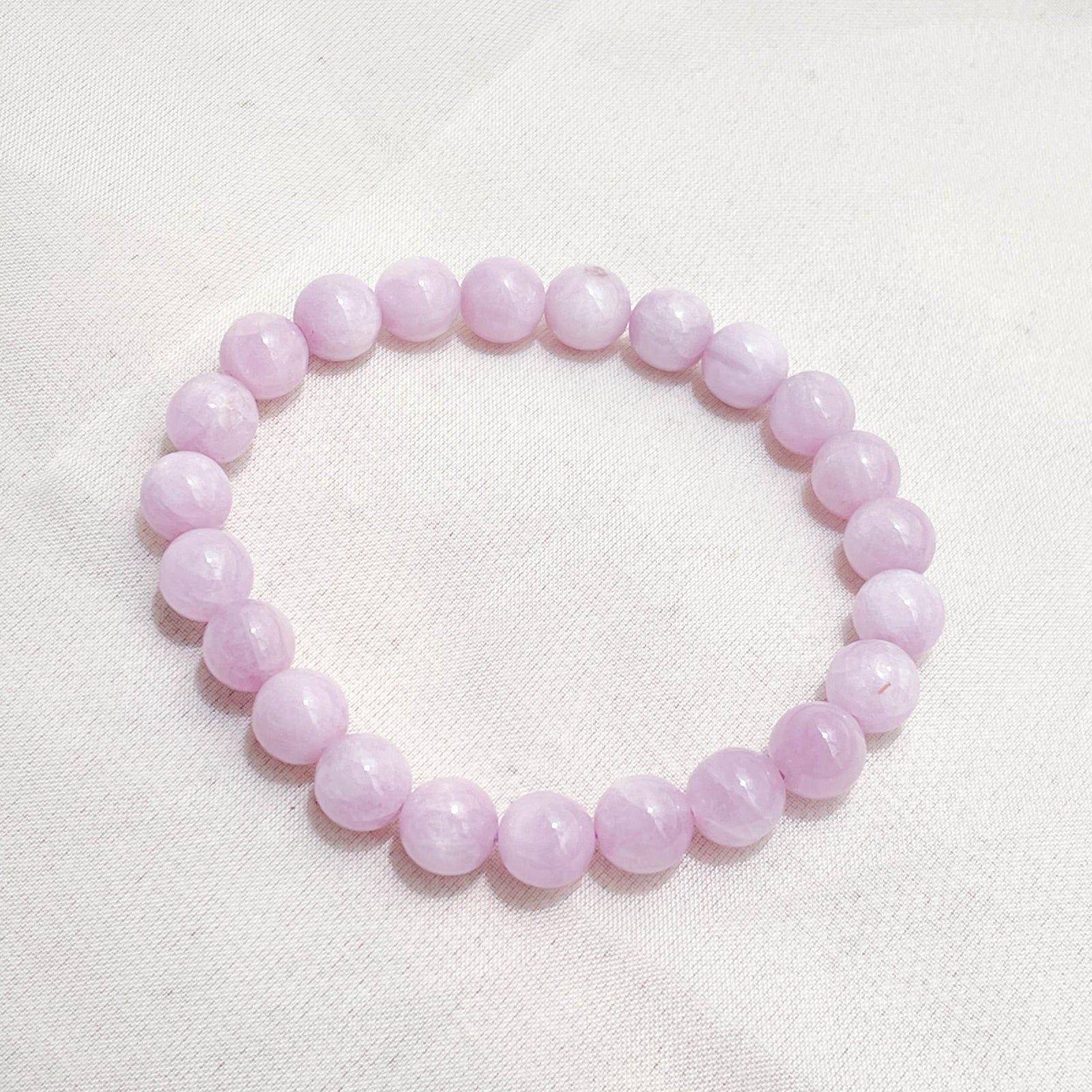 The Healing Hedge Witch's Kunzite Crystal Bracelet, made of round, pale pink beads, is displayed on a light-colored, textured fabric background.