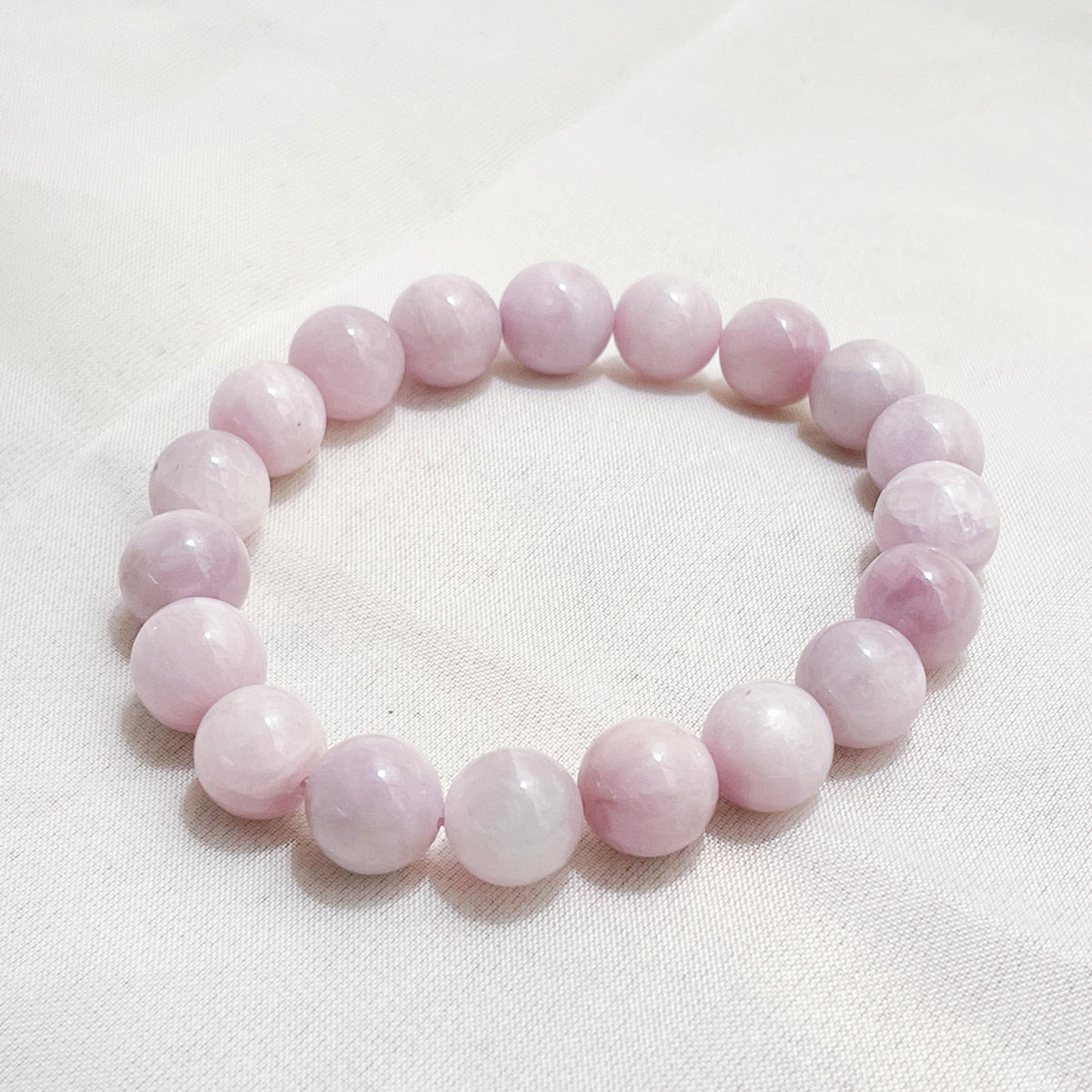 The Healing Hedge Witch’s Kunzite Crystal Bracelet, featuring smooth, light pink round beads, is displayed in a circle on a white fabric background.
