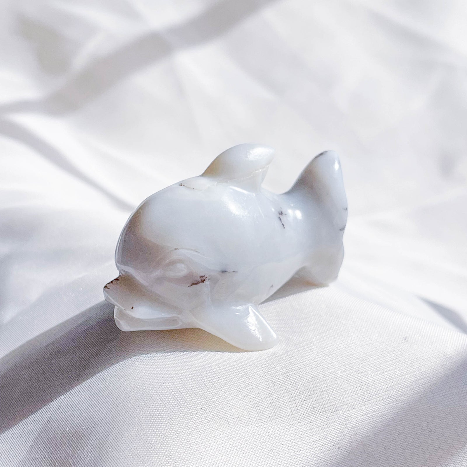 The Healing Hedge Witch’s Crystal Dolphin is a small, smooth white sculpture with gentle features and subtle grey veins, displayed on soft sunlit fabric.