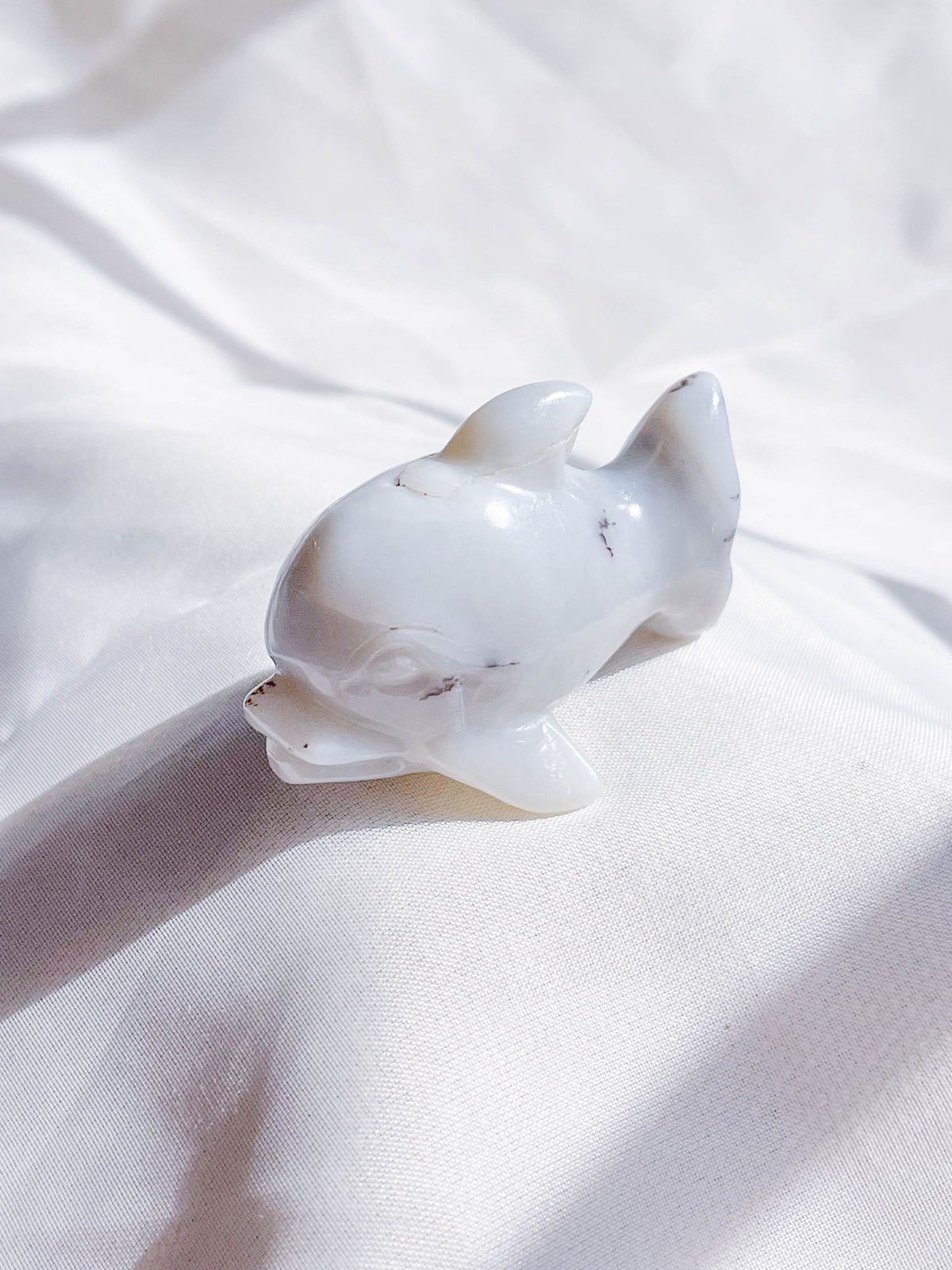 The Healing Hedge Witch's Crystal Dolphin, a smooth white stone sculpture, rests on soft white fabric with gentle shadows and highlights accentuating its graceful form.