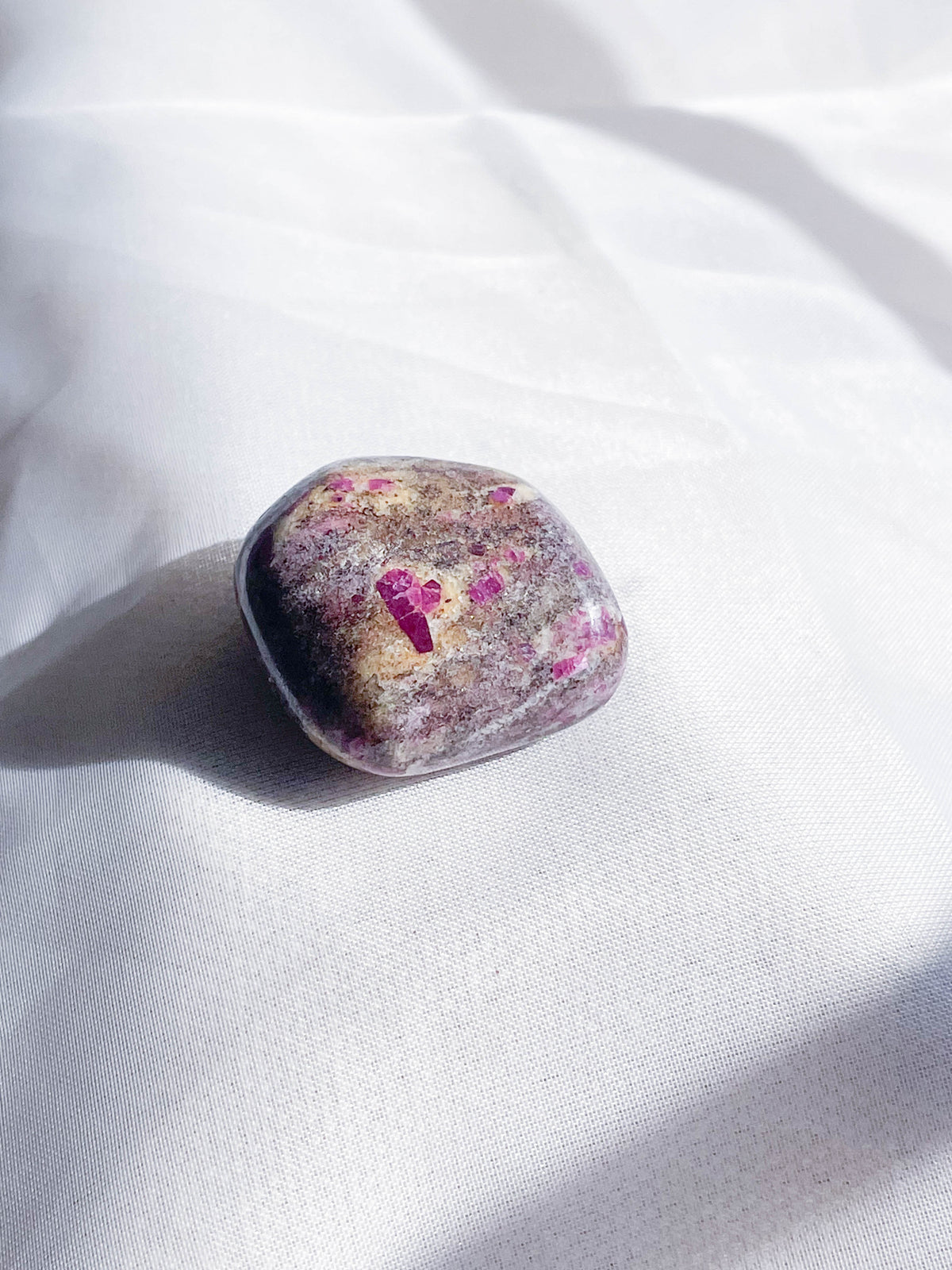 The Healing Hedge Witch's Ruby Albite Tumble, featuring purple and pink specks, rests on softly lit white fabric.