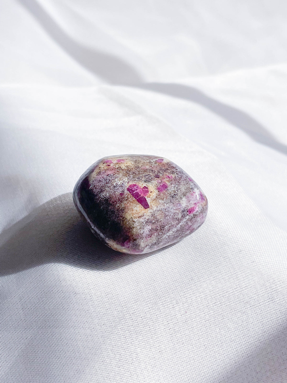 The Healing Hedge Witch Ruby Albite Tumble, an oval stone with purple and pink hues, rests on softly lit white fabric, radiating gentle Heart Chakra energy.
