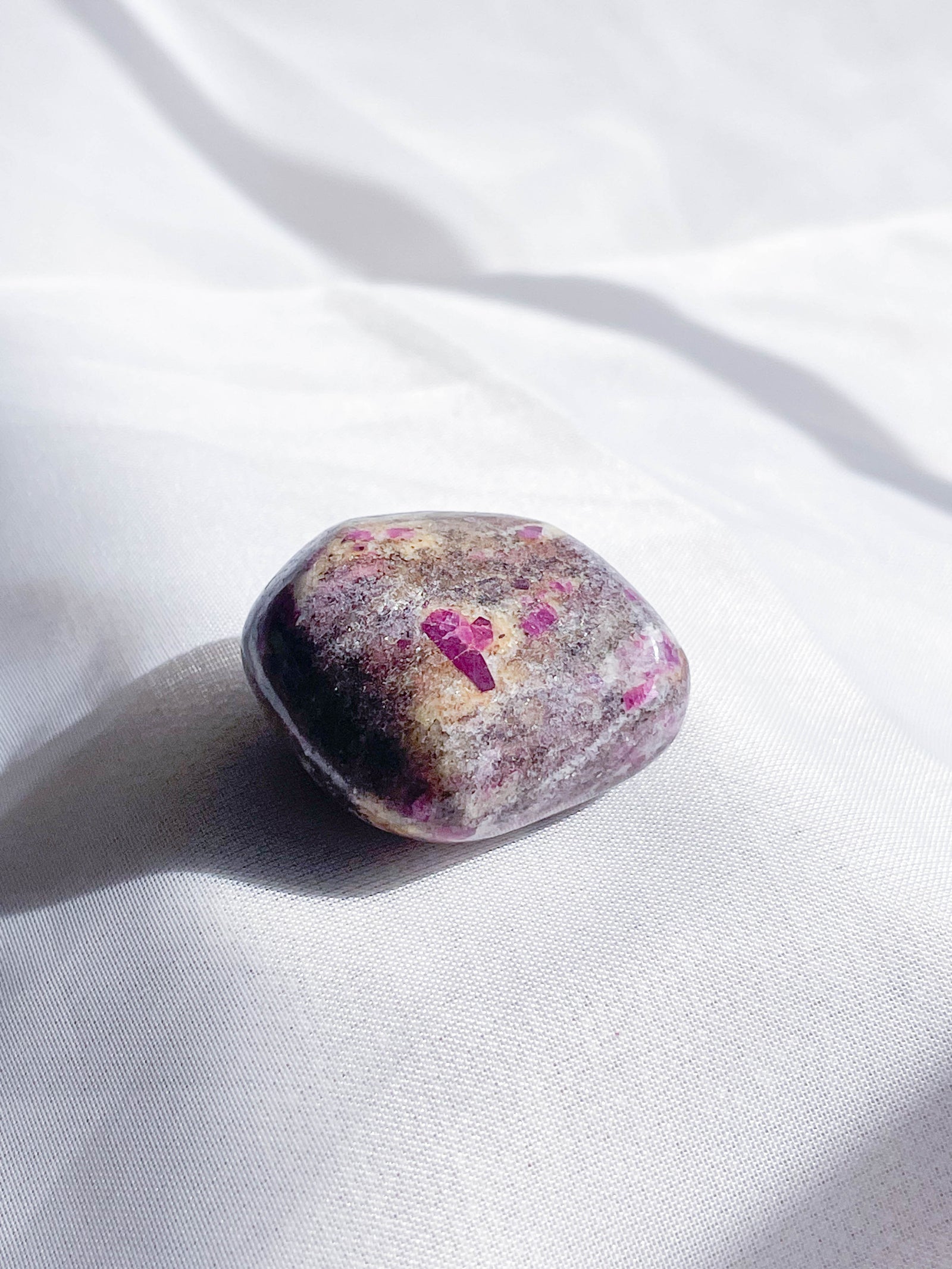 The Healing Hedge Witch Ruby Albite Tumble, an oval stone with purple and pink hues, rests on softly lit white fabric, radiating gentle Heart Chakra energy.