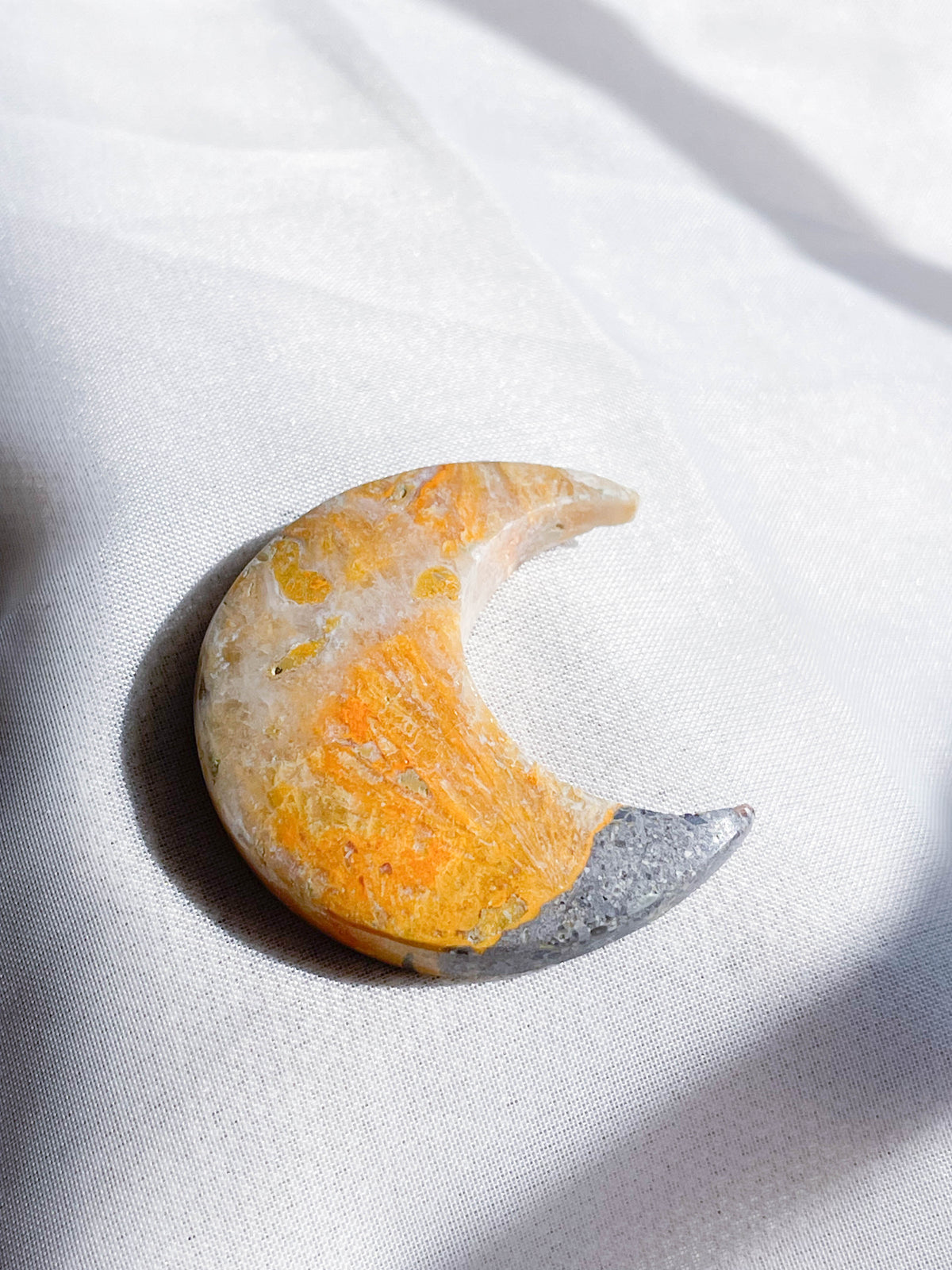 The Healing Hedge Witch's Bumblebee Bee Jasper Shape, crescent moon-shaped with yellow, orange, white, and gray hues, rests on a white fabric surface in soft natural light.