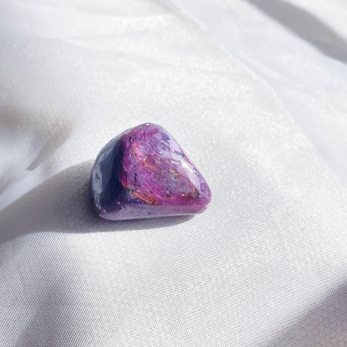 A Ruby Tumble by The Healing Hedge Witch, this polished purple gemstone with pink and red hues rests on soft white fabric, catching light—a healing stone reputed to balance chakras.