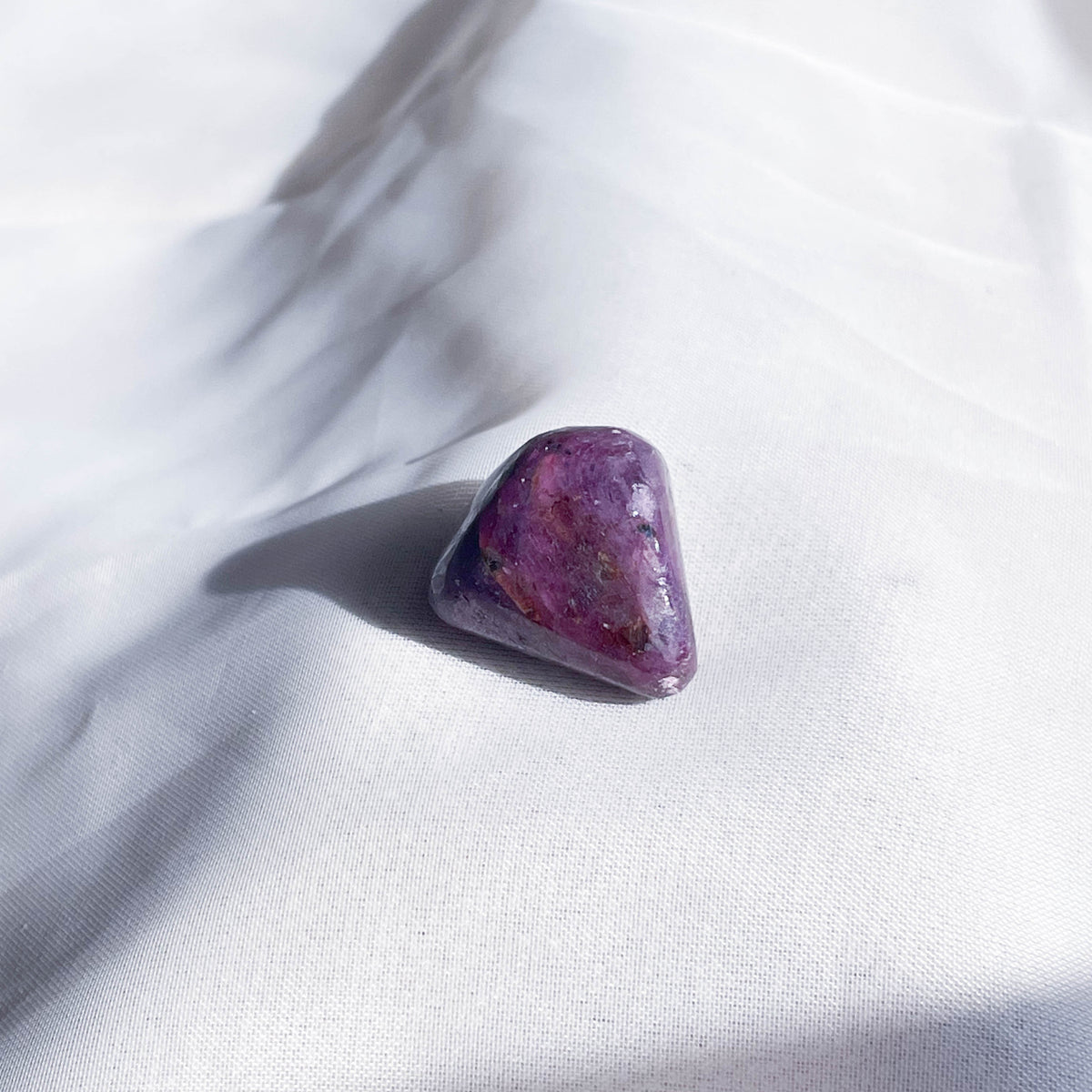 The Healing Hedge Witch’s Ruby Tumble rests on softly textured white fabric, with gentle sunlight and shadows enhancing the stone’s polished purple surface and the fabric’s delicate folds.