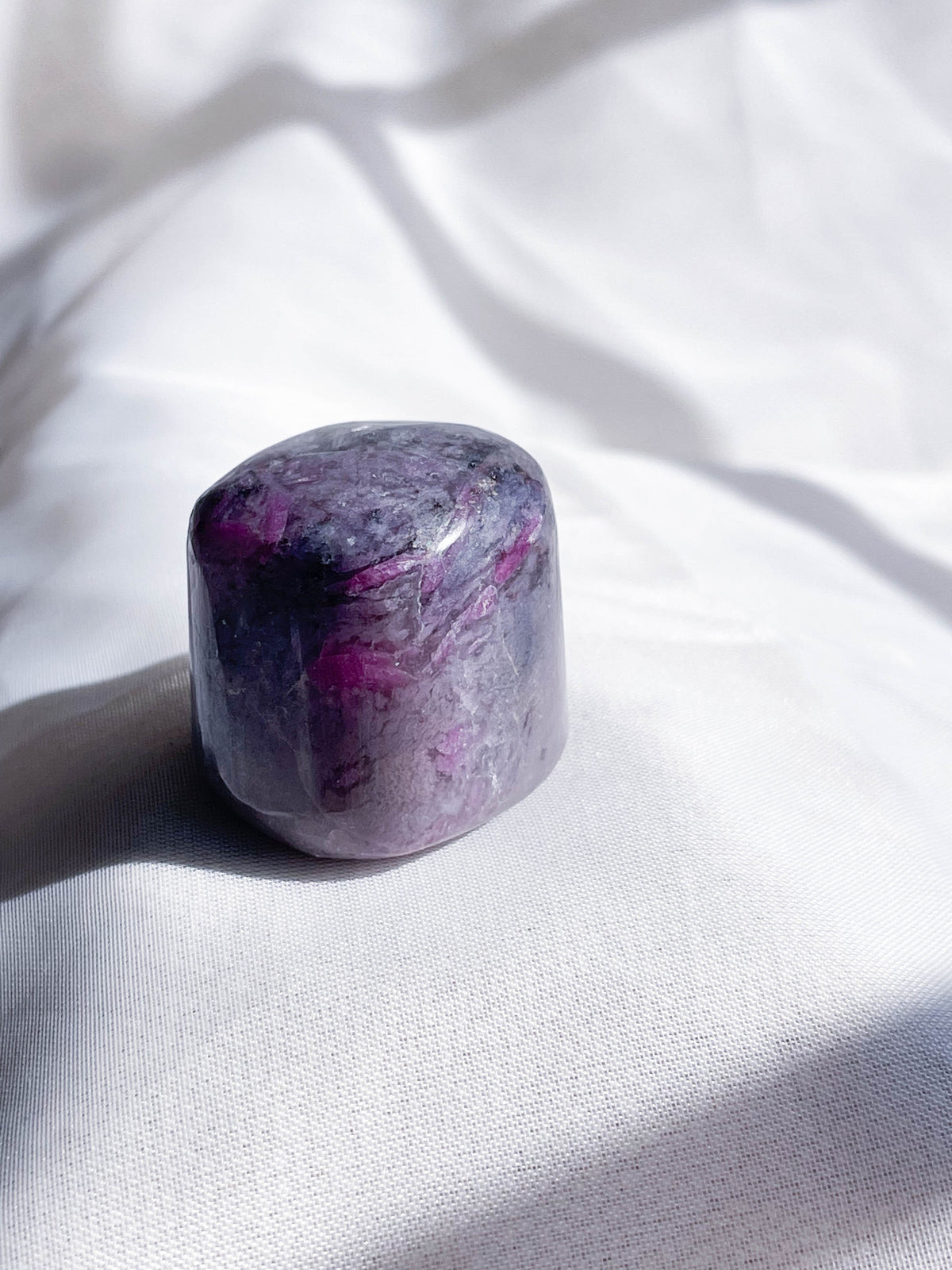 A polished, rounded Ruby Quartz Tumble by The Healing Hedge Witch—with hints of white and deep purple—rests on white fabric in natural light, making it a beautiful Heart Chakra healing stone.