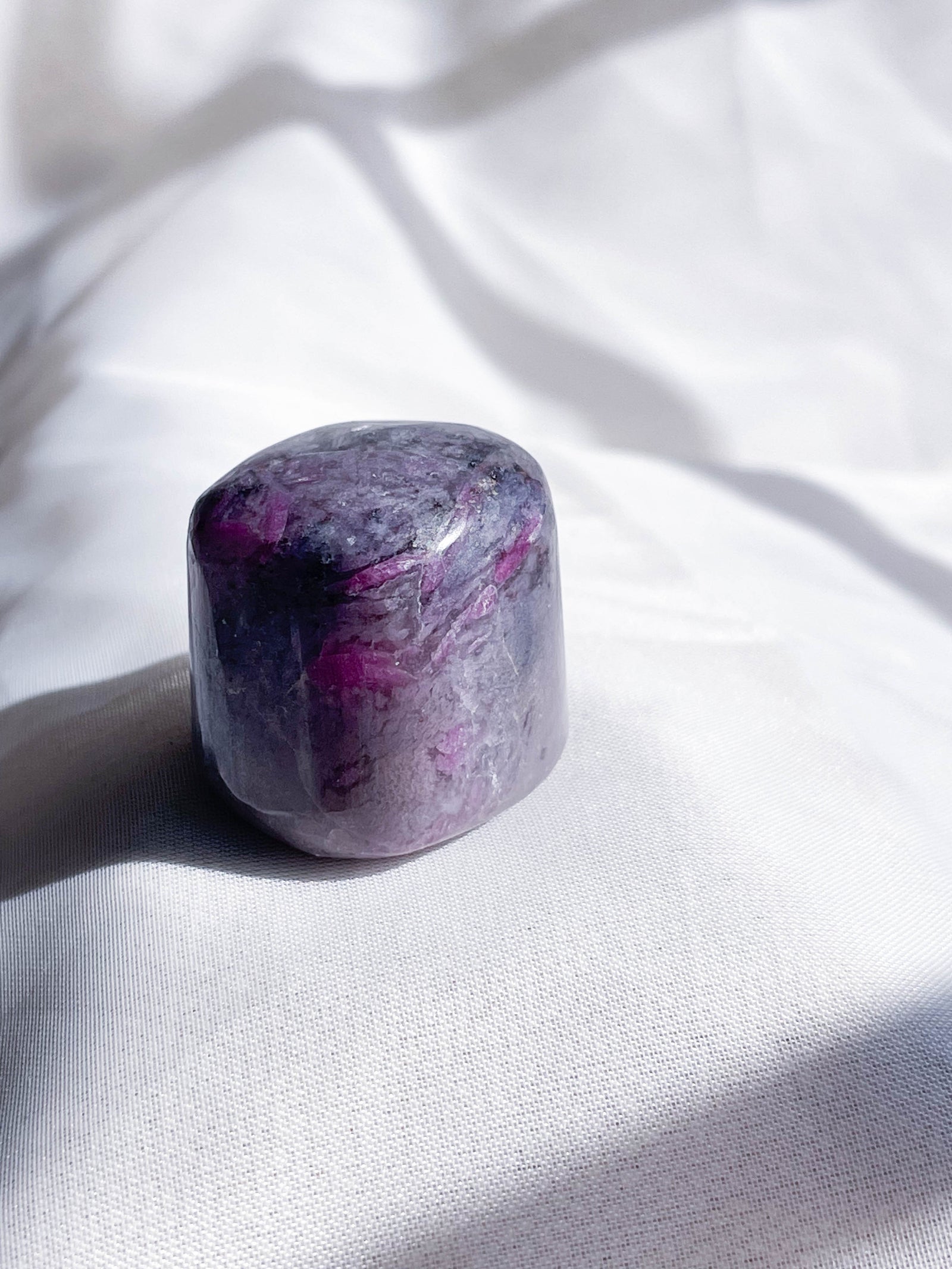 A polished, rounded Ruby Quartz Tumble by The Healing Hedge Witch—with hints of white and deep purple—rests on white fabric in natural light, making it a beautiful Heart Chakra healing stone.