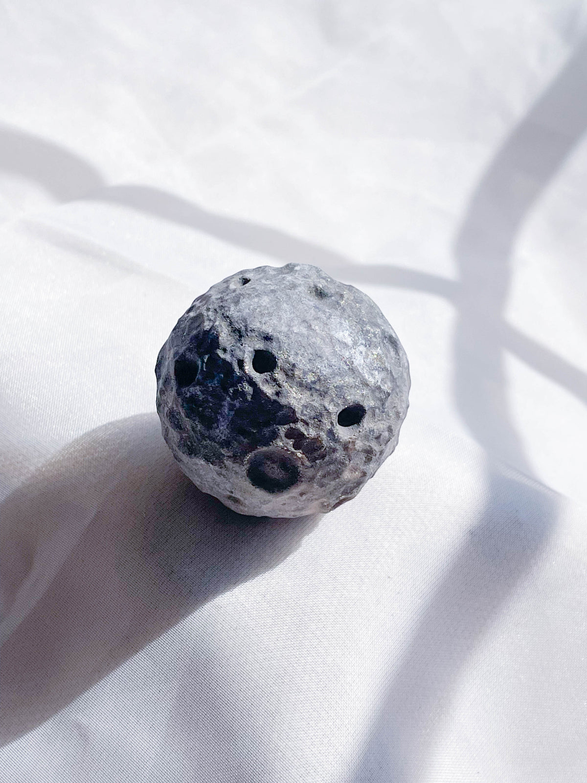 The Healing Hedge Witch’s Crater Crystal Moon, a round gray object with multiple holes and craters, rests on a white textured surface under soft lighting with gentle shadows.