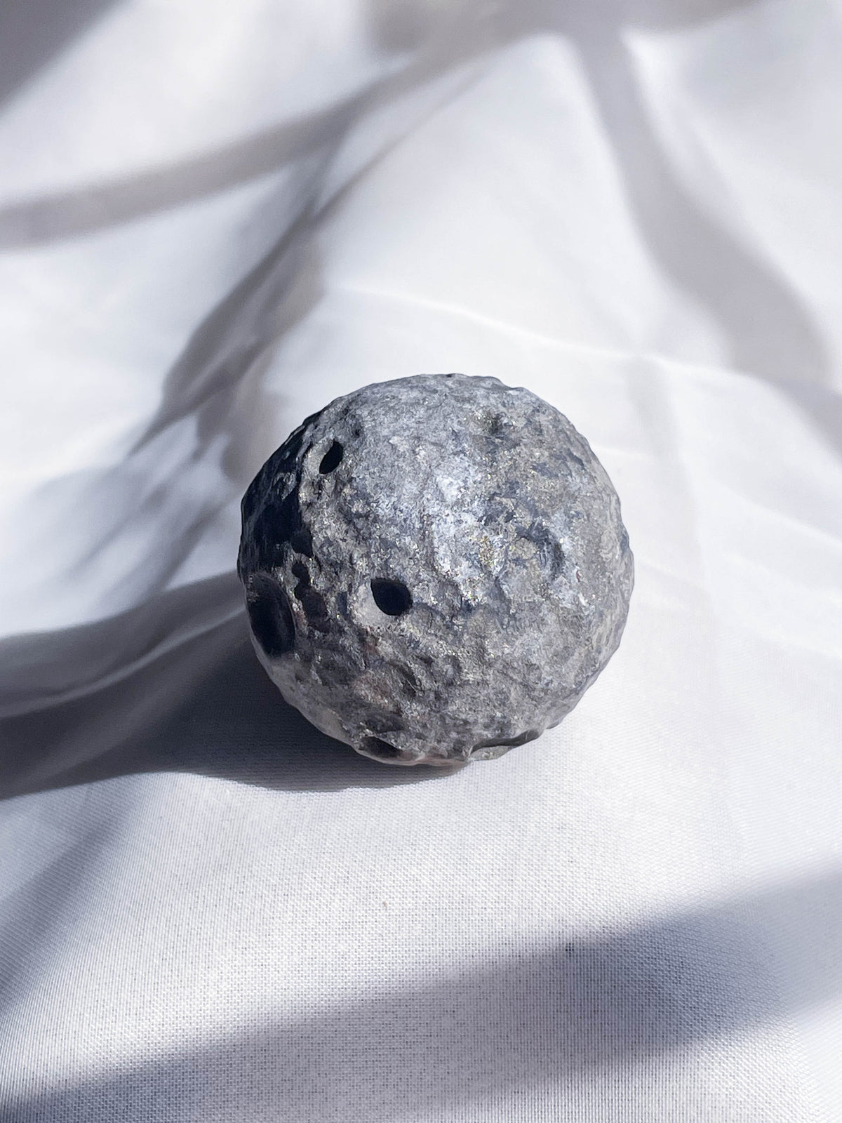 The Healing Hedge Witch’s Crater Crystal Moon—a round, gray, cratered stone—rests on softly wrinkled white fabric, its lunar look highlighted by gentle interplay of light and shadow.