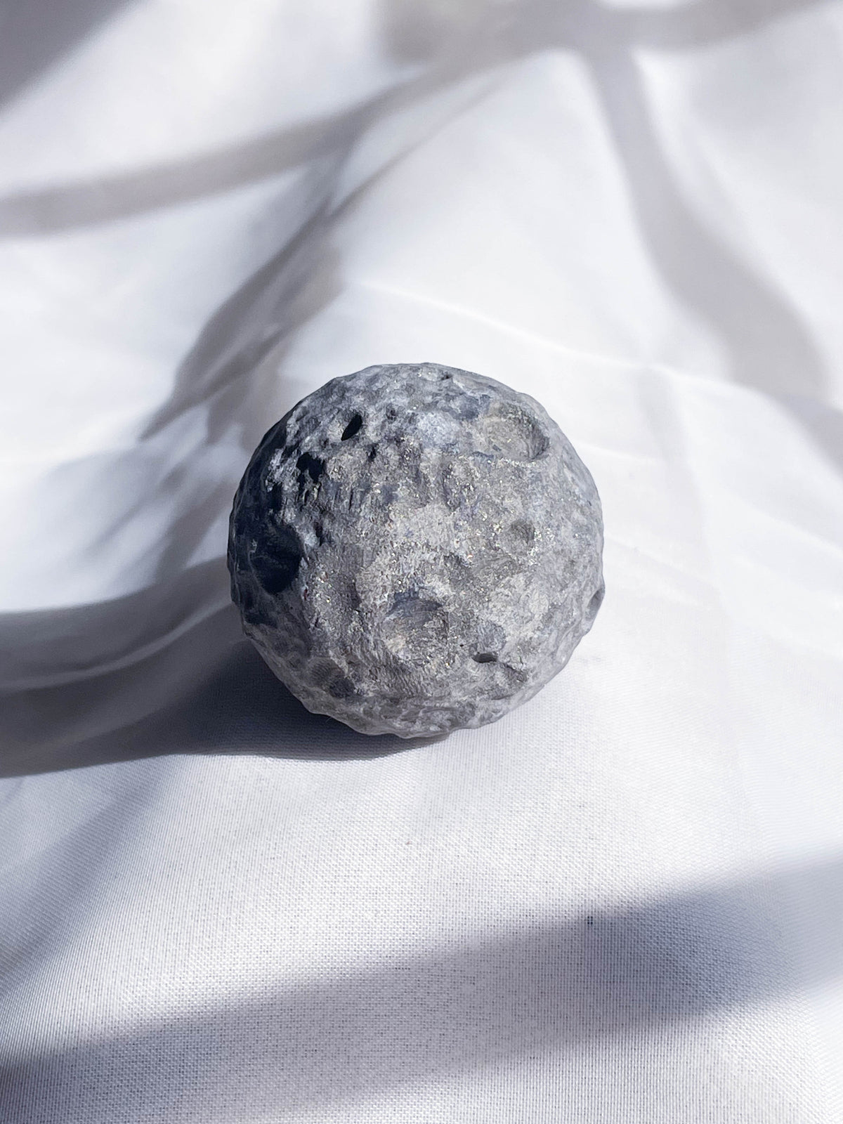 The Healing Hedge Witch's Crater Crystal Moon, a round gray stone with crater-like textures, rests on softly wrinkled white fabric, beautifully lit by natural light and gentle shadows.