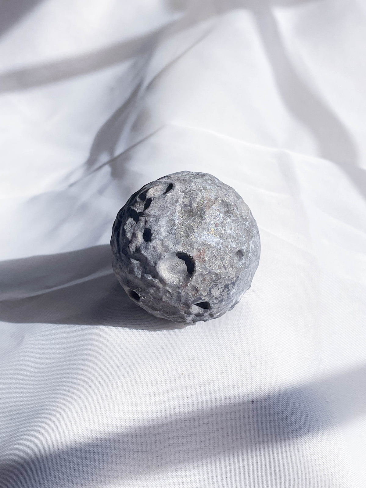 The Healing Hedge Witch’s Crater Crystal Moon, a small gray round decor piece with crater-like indentations, sits on softly wrinkled white fabric, casting a shadow and resembling a miniature moon.